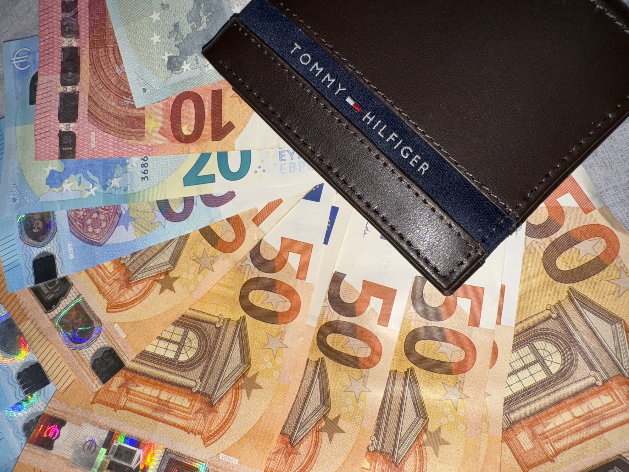 The image shows a brown leather Tommy Hilfiger wallet placed on top of a collection of Euro banknotes. The banknotes include 50 Euro notes, 20 Euro notes, 10 Euro notes, and 5 Euro notes. The location is Unknown, Unknown.