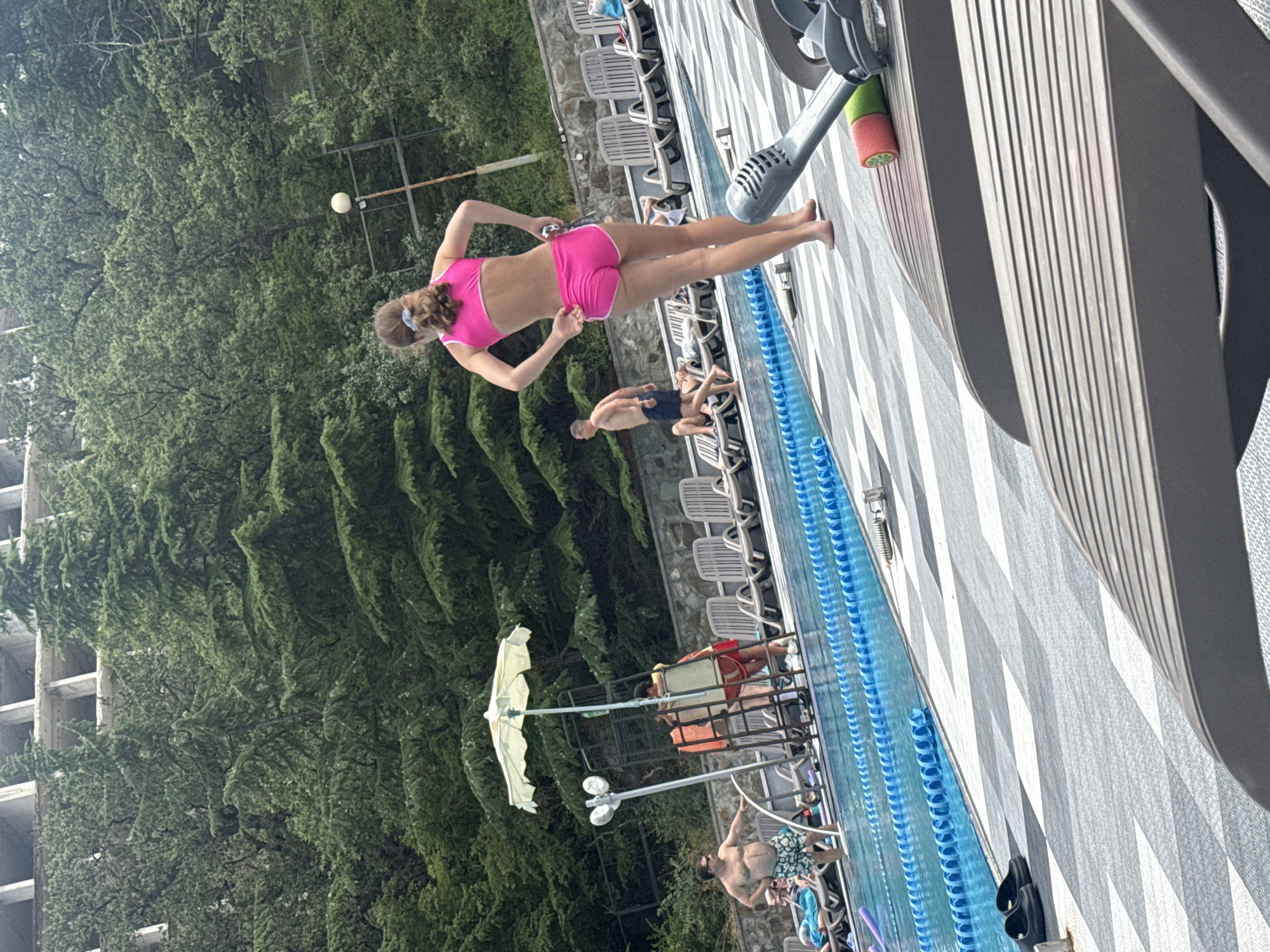 A young woman in a bright pink bikini set stands on a patterned poolside patio, facing away from the camera, with her hands on her hips. Behind her, a swimming pool with blue lane markers stretches across the scene. Several lounge chairs are visible on the patio, some occupied by people. To the left, a lifeguard stands on a raised platform under a yellow umbrella, interacting with another person. The background is dominated by lush green trees and a partially constructed building. The overall scene depicts a resort or hotel poolside environment in Массандровский поселковый совет, Ukraine, during daylight hours.