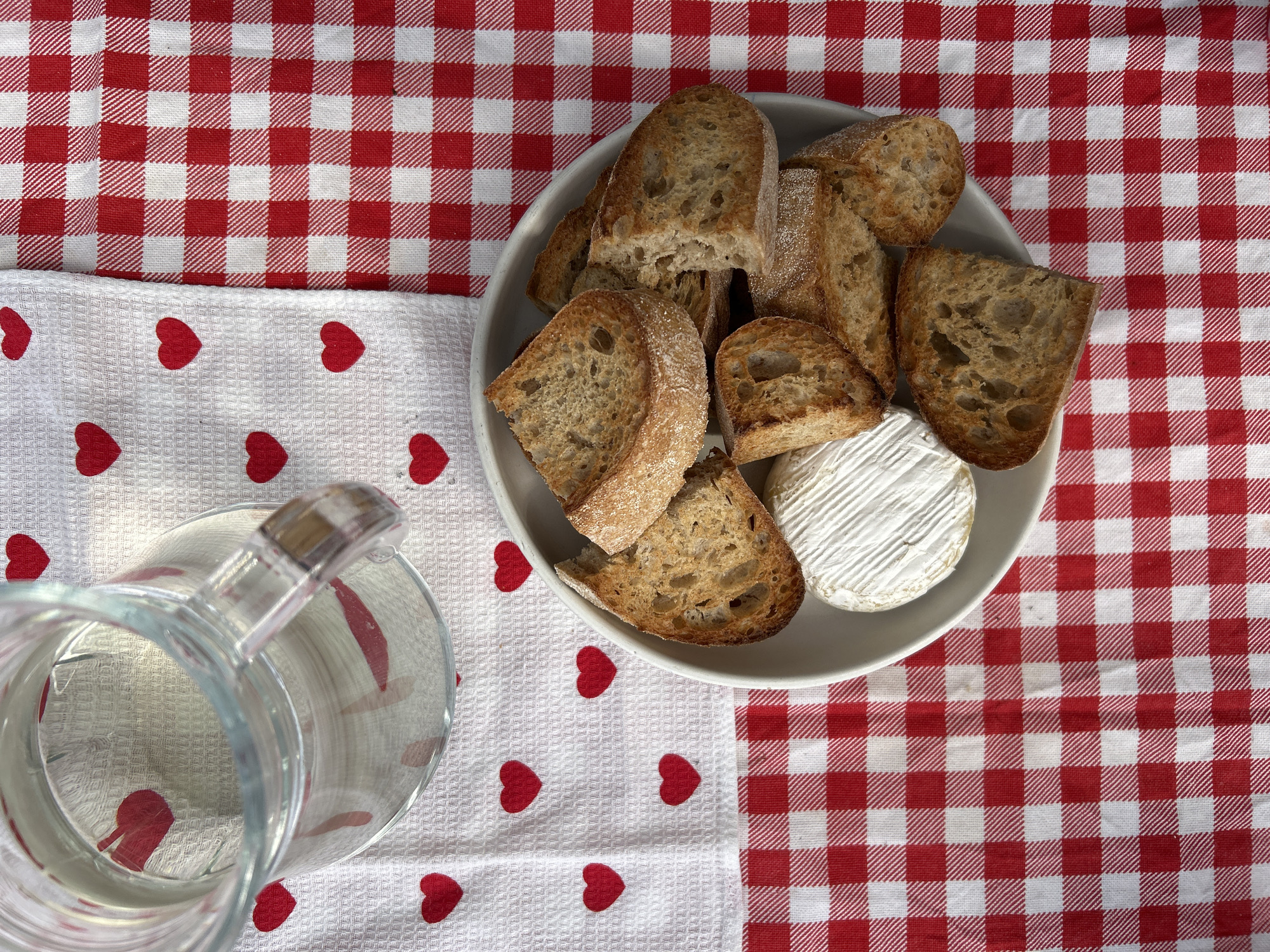 The image shows a close-up overhead shot of a rustic picnic setting. The background is a red and white checkered tablecloth. A white dish towel with red hearts printed on it partially covers the tablecloth. In the lower left corner, a clear glass pitcher, partially filled with liquid, is visible. A shallow gray bowl sits on the tablecloth and holds slices of toasted bread surrounding a small wheel of white cheese, likely Brie. The scene is brightly lit, suggesting an outdoor setting, though the location of Rautalakhti, Russia is not discernible from the image. There are no people visible.