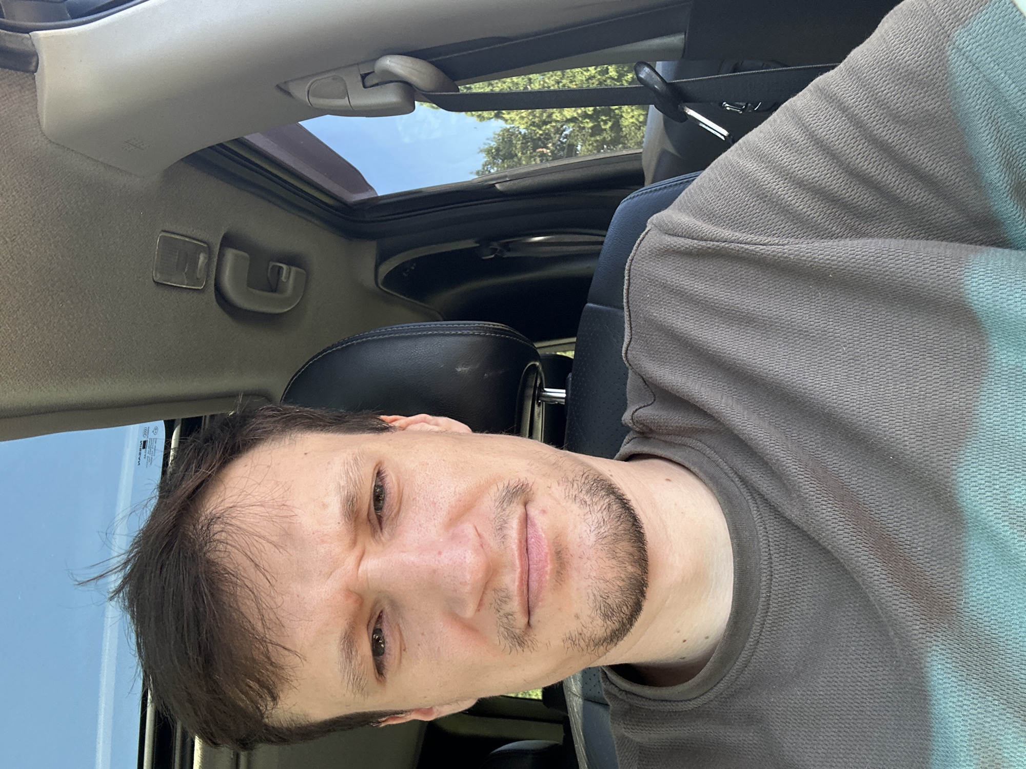 A man is seated in the driver's seat of a car, facing forward and looking at the camera. He has short, dark brown hair, brown eyes, and a faint stubble on his chin and upper lip. He is wearing a grey t-shirt with a turquoise band across the chest. The car interior is visible, including the headrest of the passenger seat, the dashboard, and a sunroof. Sunlight streams in through the sunroof and the open passenger window, illuminating the scene and casting shadows. Outside the window, green trees and a blue sky are visible. The location is Shatava, Ukraine.