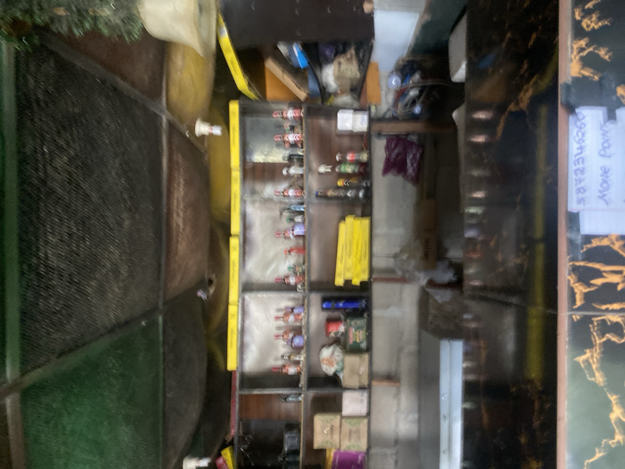 The image depicts the interior of a small establishment, possibly a shop or bar, located in Jalingo, Nigeria. The setting appears somewhat cluttered and informal. In the foreground, a dark, reflective surface, possibly a countertop or table, dominates the lower portion of the image. This surface has a marbled pattern with golden-yellow veining. A white piece of paper with handwritten text in blue ink is visible on this surface. The text is partially obscured but appears to read "5872346260" and "Move Point."
Behind the counter, shelves are visible, stocked with various items. On the upper shelves, numerous bottles, likely alcoholic beverages, are arranged. These bottles vary in size, shape, and color, with some appearing to be clear, red, and amber. Some bottles have decorative labels. On the lower shelves, there are boxes, some appearing to be cardboard or paper, along with a red cylindrical object and a blue object. A stack of bright yellow items, possibly packaging or merchandise, is also present on the lower right.
The walls and ceiling are visible, with the ceiling appearing to be constructed from a lattice-like material with some areas of green and brown discoloration. Lighting fixtures are present on the ceiling. The overall impression is of a functional, albeit somewhat disorganized, retail or service space in Jalingo. There are no people directly visible in the image.