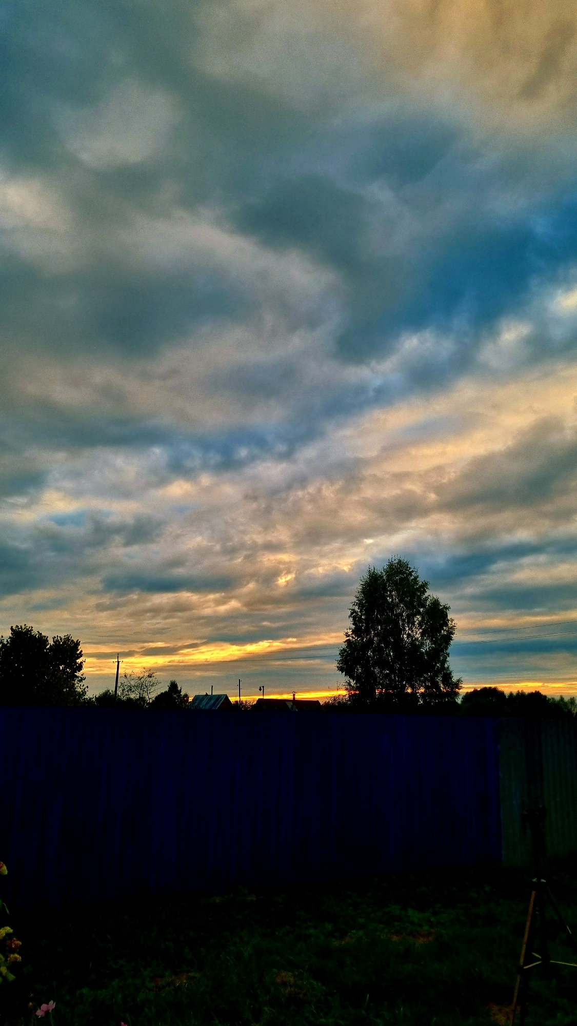 The image depicts a twilight scene over the city of Vysokoe, Russia. A dark blue fence dominates the foreground, with a patch of dark grass visible in the lower portion of the frame. Above the fence, silhouetted trees and the roofs of several houses are visible against a dramatic sky. The sky itself is filled with a mix of dark gray, blue, and yellow clouds, suggesting either an approaching storm or the aftermath of a sunset. The overall mood is serene and slightly melancholic, characteristic of a rural evening. There are no people or animals visible in the image.