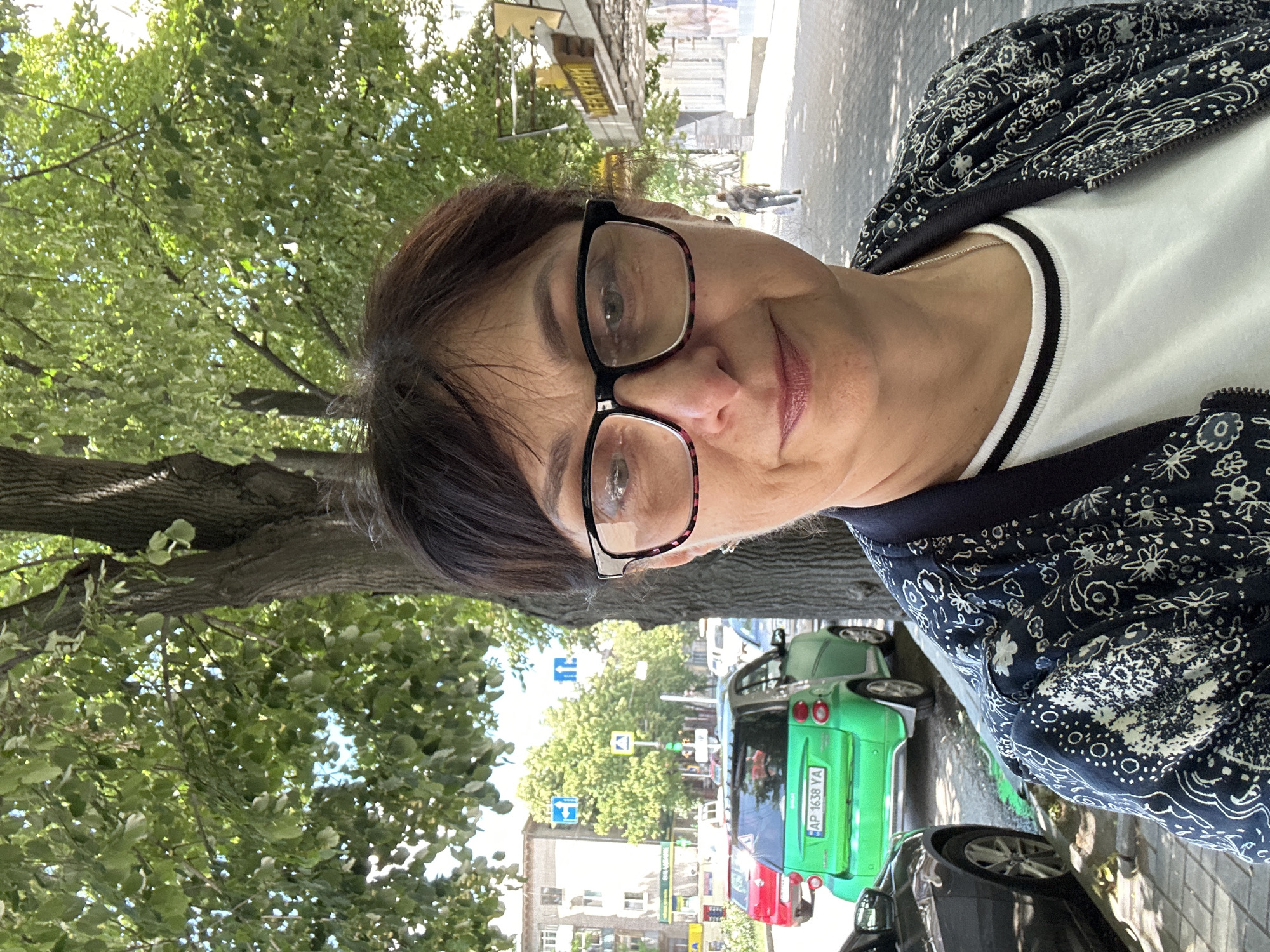 A mid-shot, outdoor, selfie-style photograph captures a middle-aged woman wearing eyeglasses and a patterned jacket over a white t-shirt with a black and white striped collar. She is standing on a city sidewalk with a large tree to her right. In the background, a street is visible with several cars, including a bright green smart car with Ukrainian license plate "AP 1638 YA", and buildings. The street appears to be in a commercial area with signage. A person is walking in the distance down the sidewalk. The scene is consistent with a street in the city of Zaporizhzhia, Ukraine, given the style of cars and signage. The lighting suggests it is daytime.