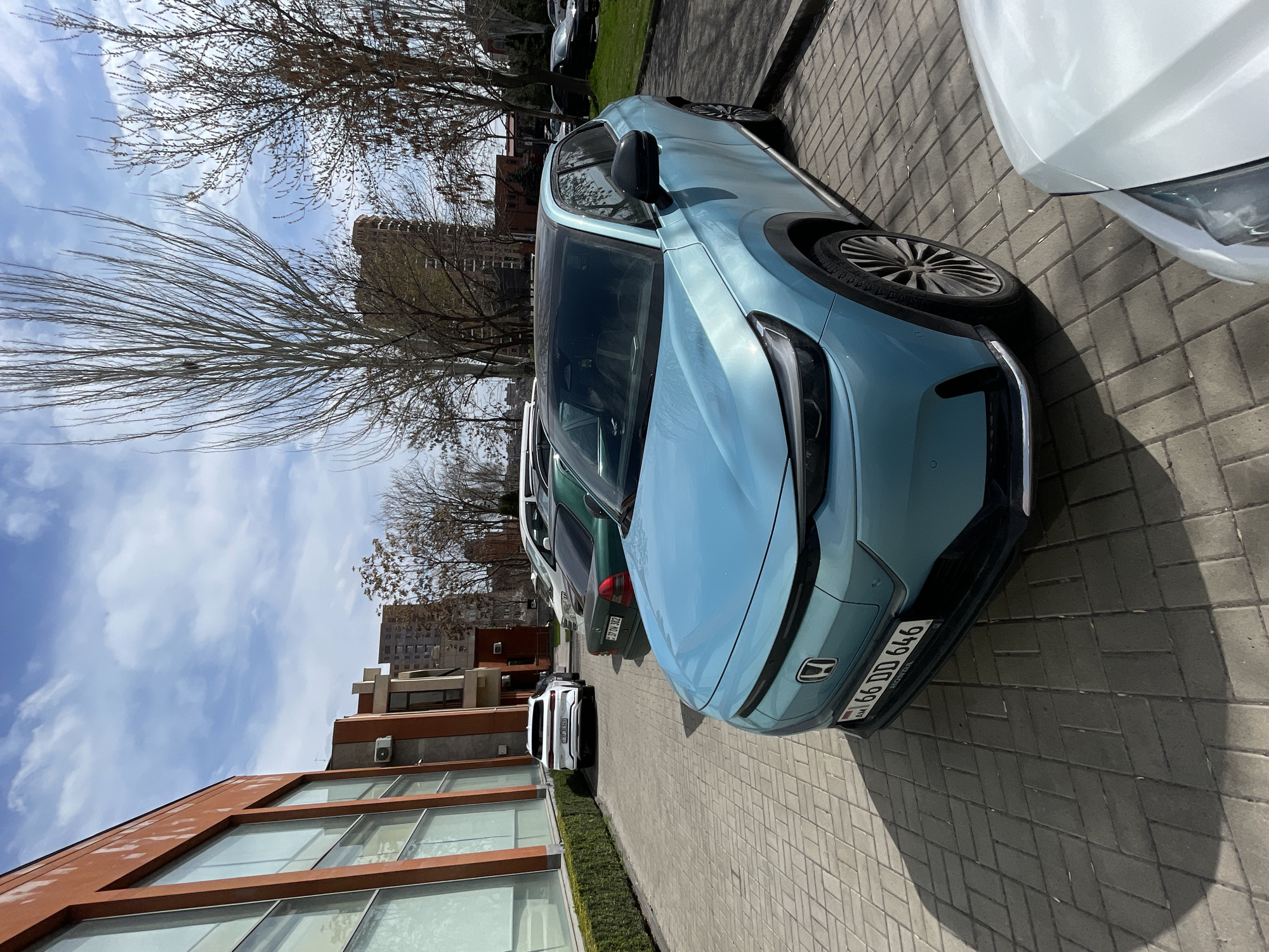 The image shows a light blue Honda parked on a brick sidewalk in Yerevan, Armenia. The license plate is visible and reads "66 DD 646." Several other vehicles are parked nearby, including a white car, a green car, and a white SUV. The background includes a multi-story building with red-brown trim and large windows, as well as bare trees and a building with a light-colored facade. The sky is partly cloudy.