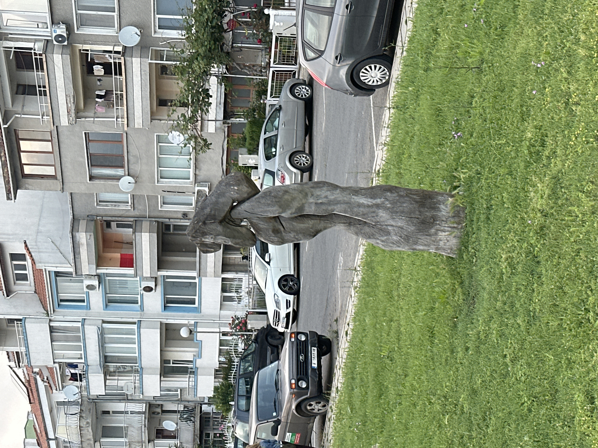 The image depicts an urban street scene in Burgas Province, Bulgaria, identifiable by the "A" prefix on the license plate. The scene features a row of multi-story residential apartment buildings in the background, a street with parked cars in the midground, and a grassy verge with a wooden sculpture in the foreground.
On the far left, a person, indistinct beyond a light-colored top, stands next to a dark gray Lada Niva SUV, which bears the Bulgarian license plate "A 1618 HK" and is parked on the street. Behind the Lada, a black SUV is parked. Further down the street, a white Mercedes-Benz C-Class sedan with dark multi-spoke wheels is parked, facing right. Next to it, a silver hatchback (possibly an Opel Astra) is parked. On the far right, partially obscured, is the rear of a dark gray hatchback.
The street is paved with asphalt and has visible parking lines. Between the street and the buildings lies a grassy verge, showing some overgrown areas and sporadic small purple and yellow wildflowers. In the foreground, on this grassy verge, a tall, weathered wooden sculpture stands upright. It is abstract, appearing to represent a stylized human form with a distinct head and torso, carved from a single piece of wood.
The background is dominated by residential buildings, which exhibit varied architectural styles, including some with light-colored facades and blue accents, and others with unpainted grey concrete. Balconies, some enclosed with windows and others open, are present on all buildings. Multiple satellite dishes and external air conditioning units are mounted on the building facades and railings. Red-tiled roofs are visible on most structures. Laundry is seen hanging to dry on a balcony railing of one of the grey buildings. A street light pole is visible in the middle ground near the buildings. The scene is illuminated by natural light, suggesting daytime with soft shadows, indicative of an overcast sky or late afternoon.
