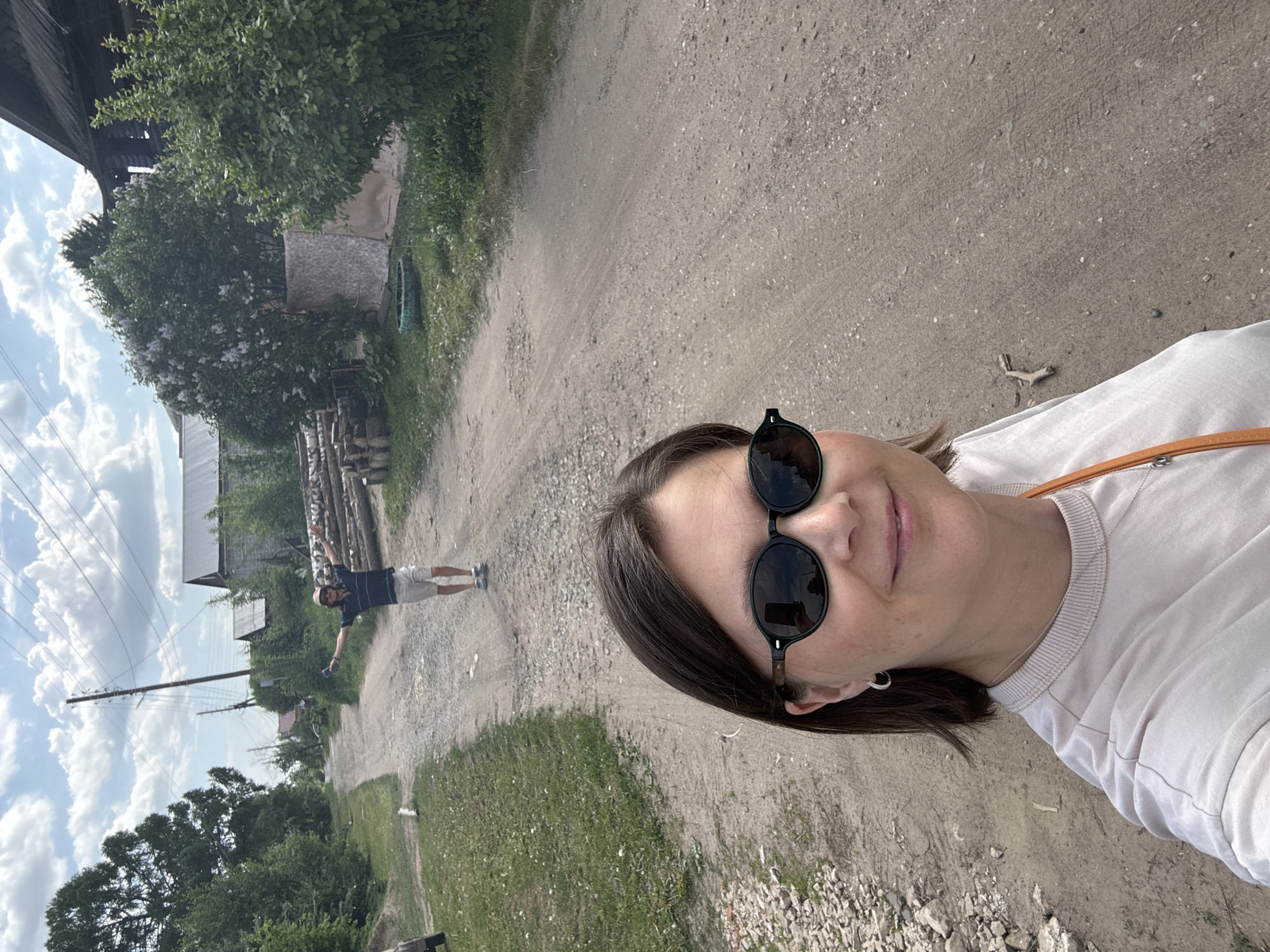 The image is a selfie taken outdoors. The photographer is a woman with dark hair and sunglasses, wearing a light-colored shirt. In the background, a dirt road stretches into the distance, flanked by greenery and some buildings. Further down the road, a man in a dark shirt and light shorts stands with his arms outstretched. The setting appears to be a rural or semi-rural area. Given the request, it's assumed this is somewhere in the city of Izhevsk, Russia. There is vegetation on either side of the road, and a power pole is visible. The sky is partly cloudy.