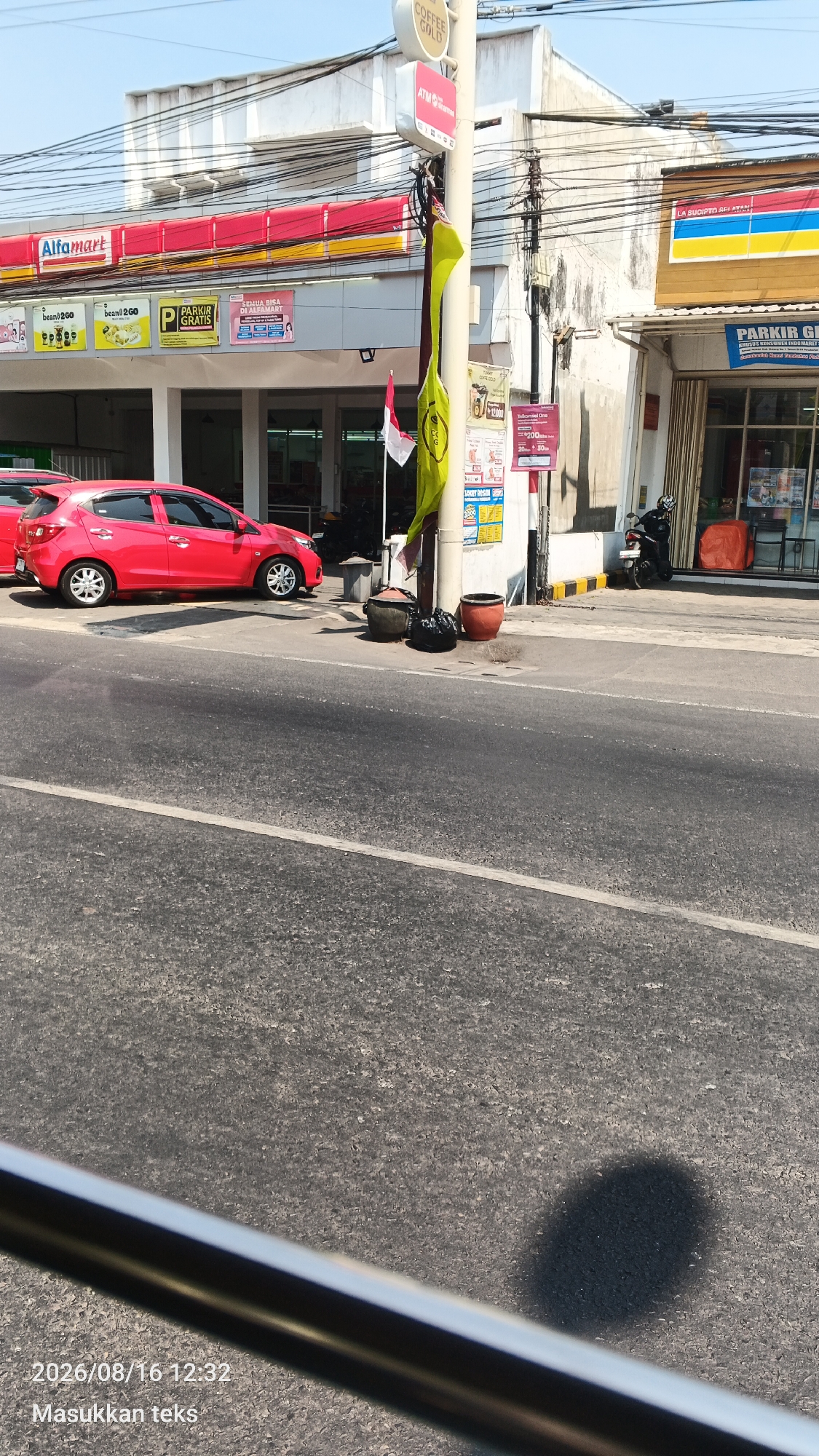 The image captures a street scene in Malang, Indonesia, featuring an Alfamart convenience store. A bright red car is parked in front of the store, partially obscuring the entrance. A street pole stands in front of the building, adorned with a yellow flag and an Indonesian flag. Several trash bins and pots are placed near the pole on the sidewalk. In the background, a motorbike is parked near another storefront. The overall environment suggests a commercial area within the city.