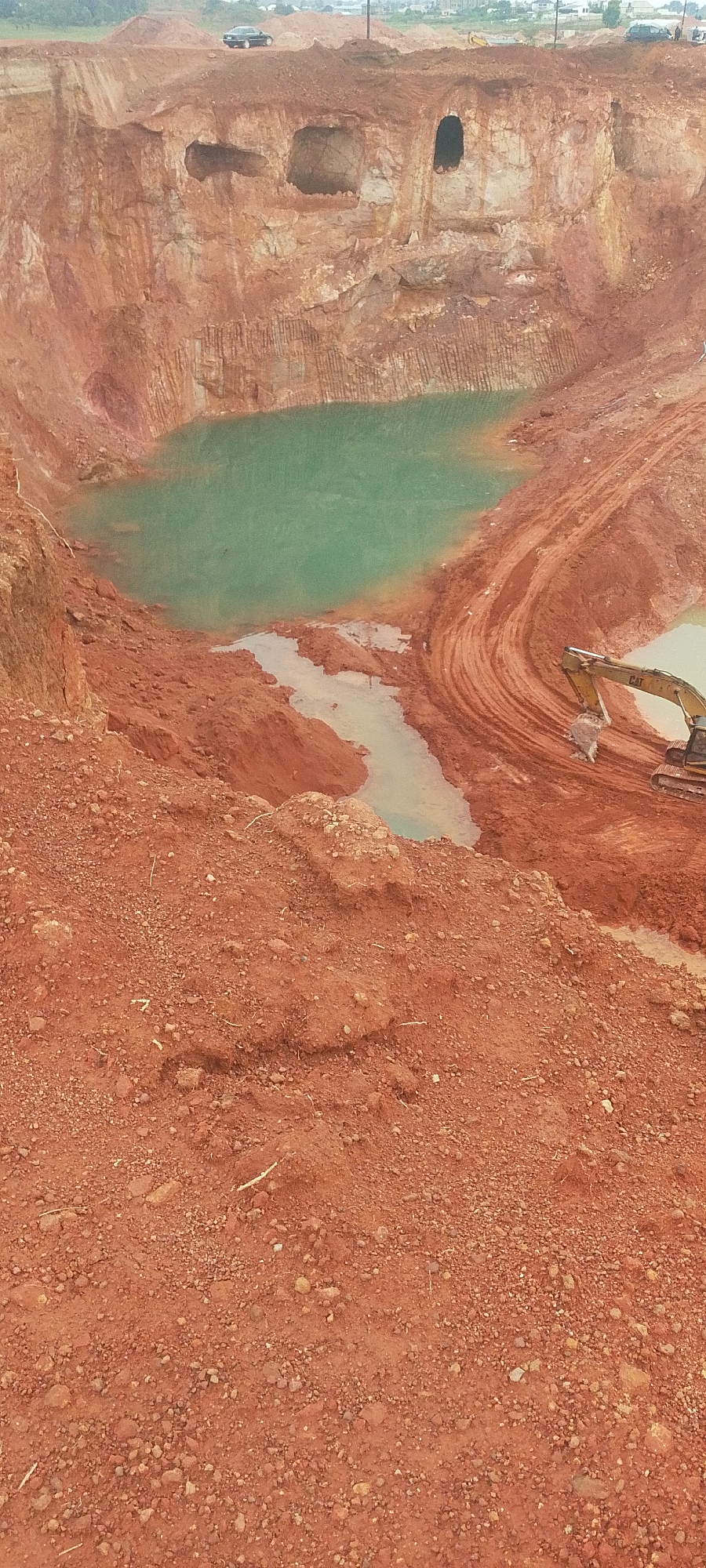 The image depicts an aerial view of a large excavation site, likely a quarry, located in Reifild-Ist, Nigeria. The dominant colors are the reddish-brown of the excavated earth and the vibrant turquoise of the water that has collected in the bottom of the pit. The sheer walls of the excavation are visible, with distinct layers and some evidence of vertical scoring from machinery. There are several dark openings, resembling caves or tunnels, carved into the upper portions of the pit walls.
In the mid-ground, a yellow excavator with the "CAT" logo is positioned on the muddy, excavated earth, its bucket lowered as if actively working. Tire tracks are visible in the mud, indicating recent activity. A small body of water, less turquoise than the main pool, is also present in this area, connected to the larger pool by a thin channel of mud and water.
In the background, atop the excavation, a dark-colored car is parked. Beyond the immediate site, the landscape suggests a transition to more developed areas, with scattered buildings and trees visible in the distance under an overcast sky. The foreground of the image is dominated by loose, reddish soil and small stones, suggesting this is the raw material being excavated or the surrounding terrain. The overall impression is one of ongoing industrial activity in a rugged, earthy environment.