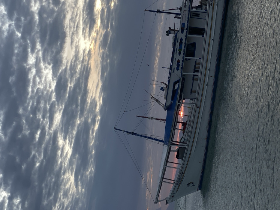 The scene is set in Juan Griego, Venezuela, at dusk or dawn. A white and blue fishing vessel, identified by "NATHALIA" on its bow and "MARIA - CARMEN" on its side, is anchored or moored in calm, dark water. The boat features a covered deck, multiple masts with rigging, and a cabin section adorned with blue and white decorative patterns. A black marlin or sailfish emblem is painted on the bow. The sky is heavily overcast with altocumulus clouds, revealing a warm, orange-pink sun near the horizon. Distant dark hills or mountains are visible on the horizon line behind the vessel. A single dark bird is captured in flight above the boat. No individuals are visible.
