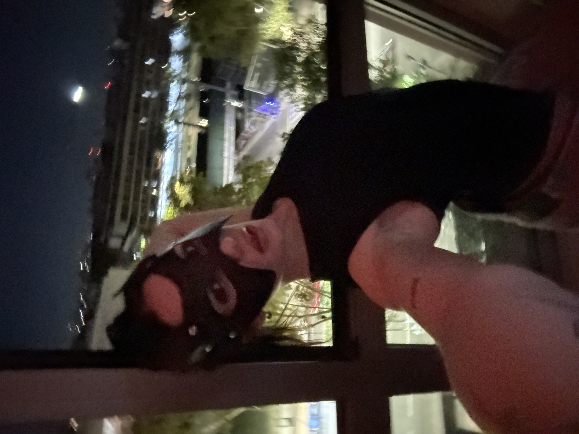 An individual is captured in an urban night scene, posing for the camera from what appears to be a high-rise building or balcony. The image is oriented as if the person is leaning over a ledge or railing, looking upwards directly at the viewer. They are wearing a distinctive black mask that covers most of their face, featuring ear-like protrusions and pinkish-red accents around the eye and mouth openings. Dark hair is visible around the mask. The person is dressed in a dark, short-sleeved top.
In the background, visible through a window or from a balcony, is a dark night sky. A prominent crescent moon glows in the upper left quadrant. Below the moon and extending across the background are the illuminated windows of numerous tall city buildings, suggesting a dense urban environment. Patches of trees and foliage, subtly lit by the city's ambient light, are also present. The scene depicts a clear, nighttime atmosphere in a bustling city. Based on the provided context, the location is Buenos Aires, Argentina. There is no visible text in the image.