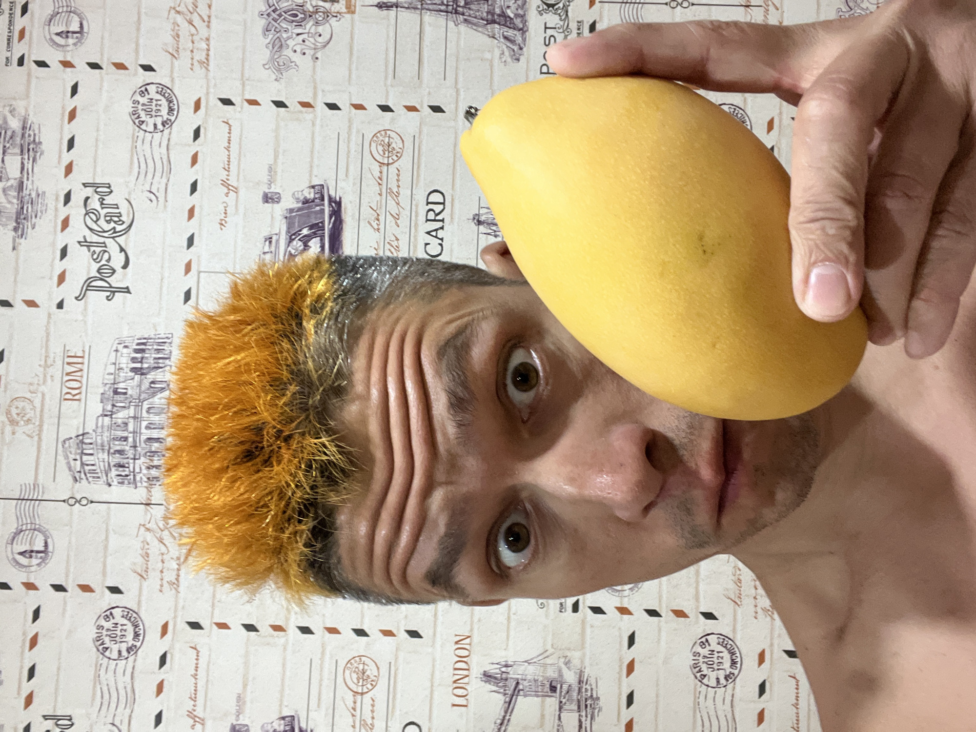 A man with orange hair holds a yellow mango in front of his face. The background is a wall with a postcard pattern featuring cities such as London, Paris, and Rome. It is unlikely this photo was taken in the city of Chaikovsky, Russia, but rather appears to be taken indoors against a patterned wall. The man looks directly at the camera.