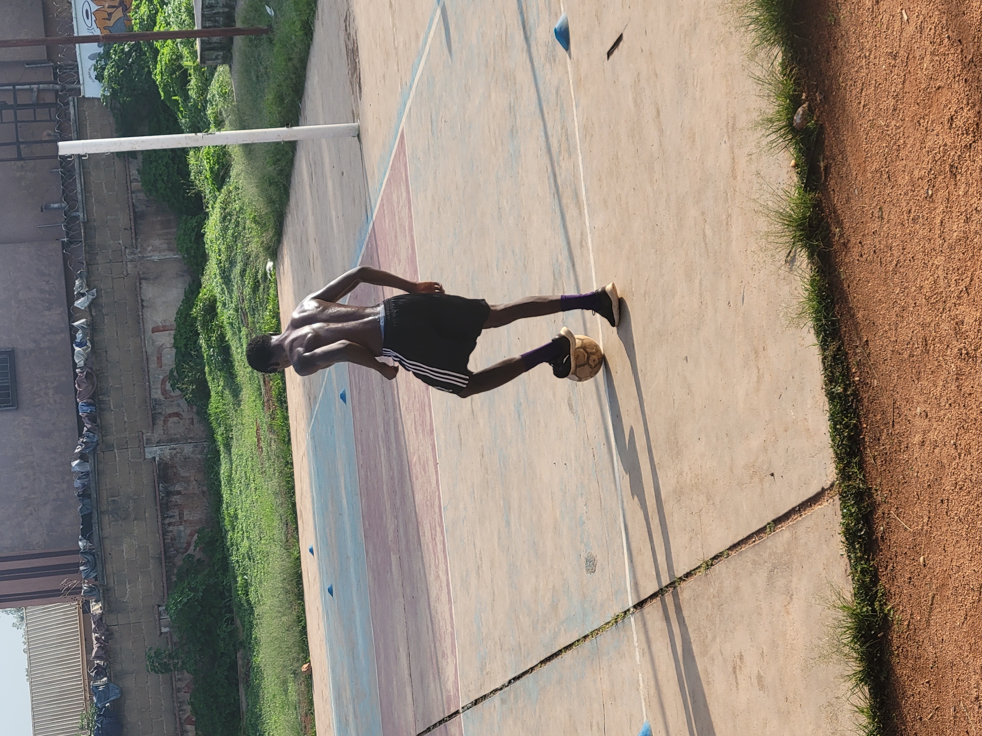 A shirtless Black male is dribbling a soccer ball on a multi-colored outdoor court in Jos, Nigeria. The man is wearing black shorts with white stripes and purple socks. He is right-footed and has his left foot on top of the soccer ball. The court has blue and red painted sections. In the background, there is a concrete wall with barbed wire on top, overgrown green vegetation, and a white pole. The ground is a mixture of concrete, dirt, and patches of grass. The image is taken from a slightly elevated angle.
