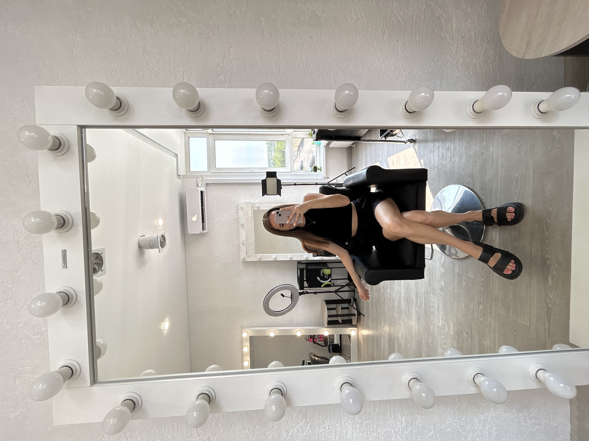 A full-length mirror with lights around its frame reflects a young woman sitting in a black chair. The woman is wearing a black crop top and black shorts, and black sandals. She is holding a phone and taking a selfie. The mirror also reflects a ring light, a makeup station, and a window with a view of the outdoors. The room appears to be a salon or beauty studio, with light-colored walls and a wooden floor. There are no identifying markers for the city or location.