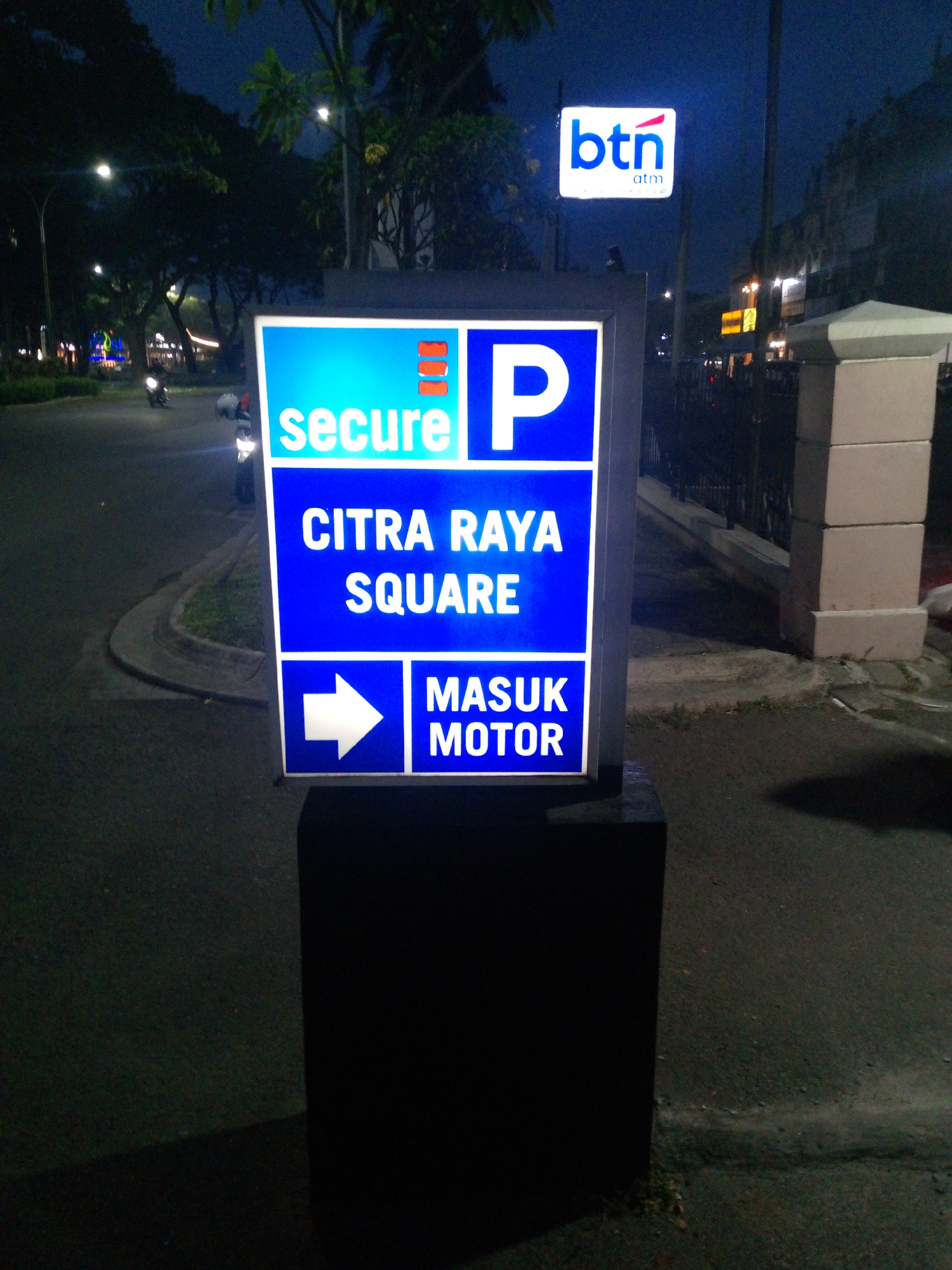 A neon-lit blue and white parking sign is the primary subject in this outdoor night scene, likely located in the city of Cikupa, Indonesia. The sign features the word "secure" in white text on the top left, and a large white letter "P" on the top right, signifying parking. Below this, the words "CITRA RAYA SQUARE" are prominently displayed in white text on a blue background. The bottom section of the sign shows a white arrow pointing right, adjacent to the text "MASUK MOTOR" (entrance for motorcycles) in white on a blue background.
In the background, a street is visible with streetlights illuminating the area, and trees line the side of the road. A motorcycle is parked near the sign. Above and to the right of the main sign, a smaller illuminated sign for "btn atm" is present. The environment appears to be a public area with paved surfaces and some landscaping. The lighting is artificial due to the signs and streetlights.