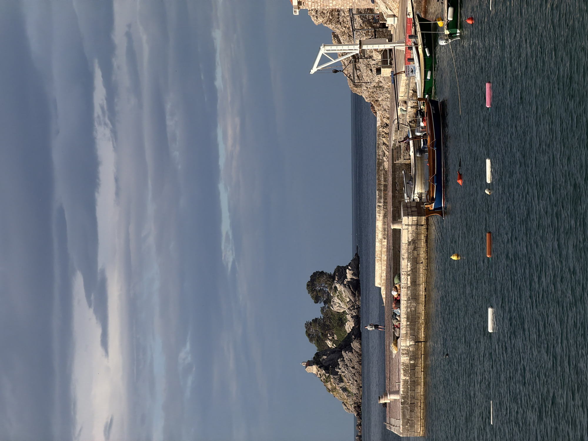The image shows a waterfront scene in Petrovac Centar, Montenegro. The composition is split between the cloudy sky and the sea. A stone pier extends into the water, with boats docked along the side. A small, rocky islet with trees and a structure on top is visible next to the pier. One person is standing on the pier fishing, with a pile of belongings or catch next to him. Buoys and other floating objects are scattered in the water. On the right, other boats are docked. A white crane structure stands out against the rock face of the harbour.