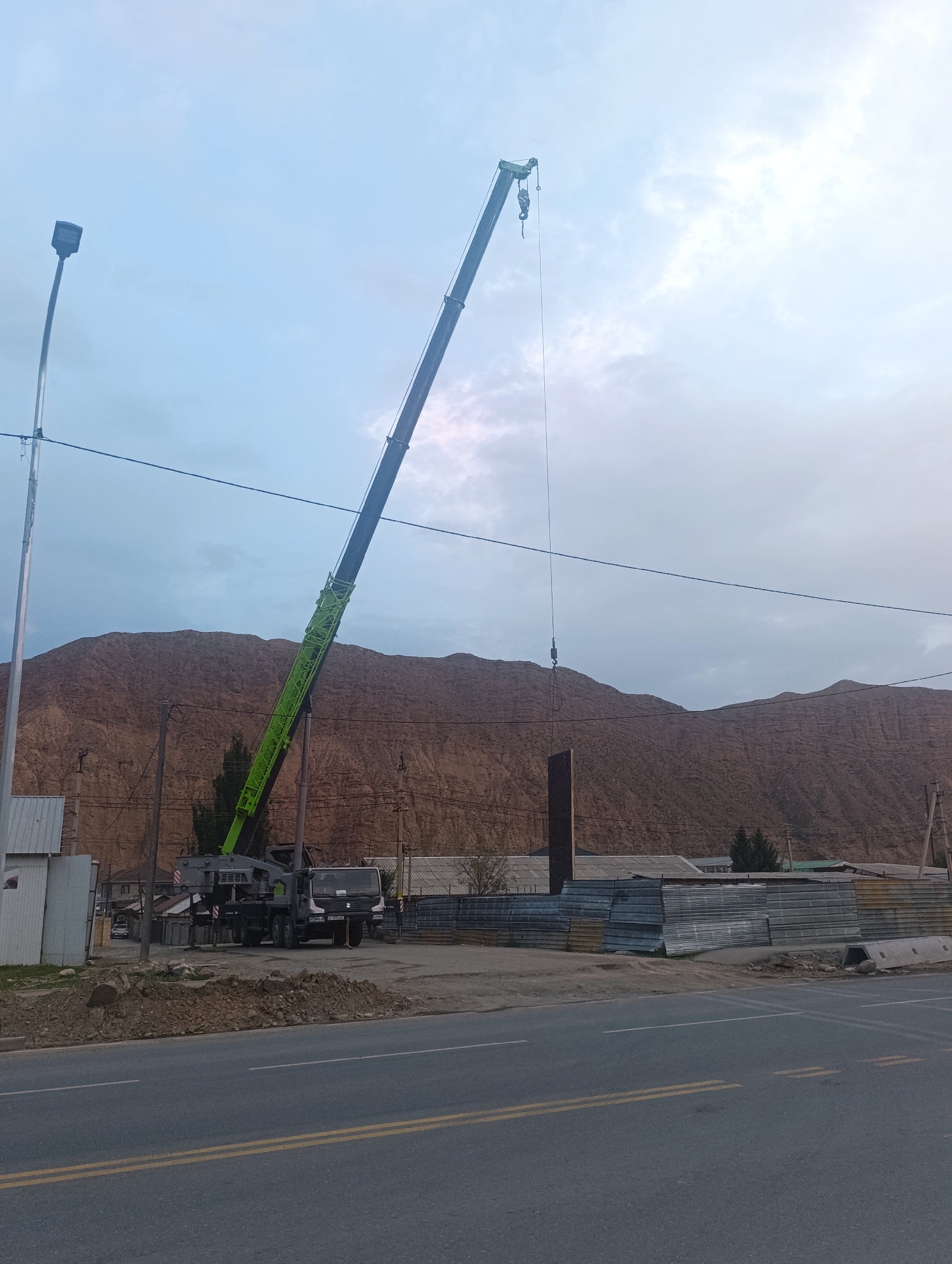 In Naryn City, Kyrgyzstan, a construction scene unfolds near a road. A large green and black crane extends its boom upwards, with a dark, vertical beam suspended from its hook. The crane is positioned behind a pile of dirt and rocks, adjacent to a gray construction truck. To the right of the truck, corrugated metal sheets are stacked. In the background, a steep, brown mountain rises under a pale, overcast sky. A streetlamp stands on the left side of the image, and utility poles with wires are visible throughout the scene. The road in the foreground has double yellow lines and a white line. The overall impression is one of ongoing construction in a semi-arid, mountainous environment.