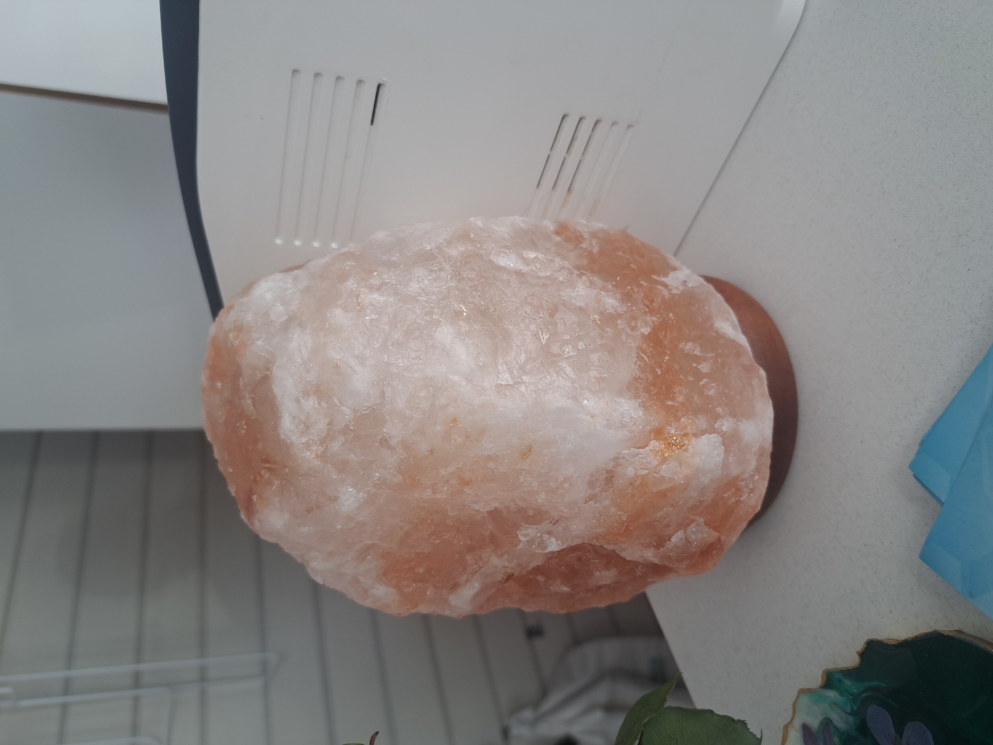A Himalayan salt lamp sits on a white surface, likely a countertop or table, partially framed by an off-white object with vertical slits. The lamp's color ranges from white to pale orange. To the lower right is a partial glimpse of a teal object and a green object. The image contains no indication of the city or location of Leningradskaya, Russia. There are no people visible.