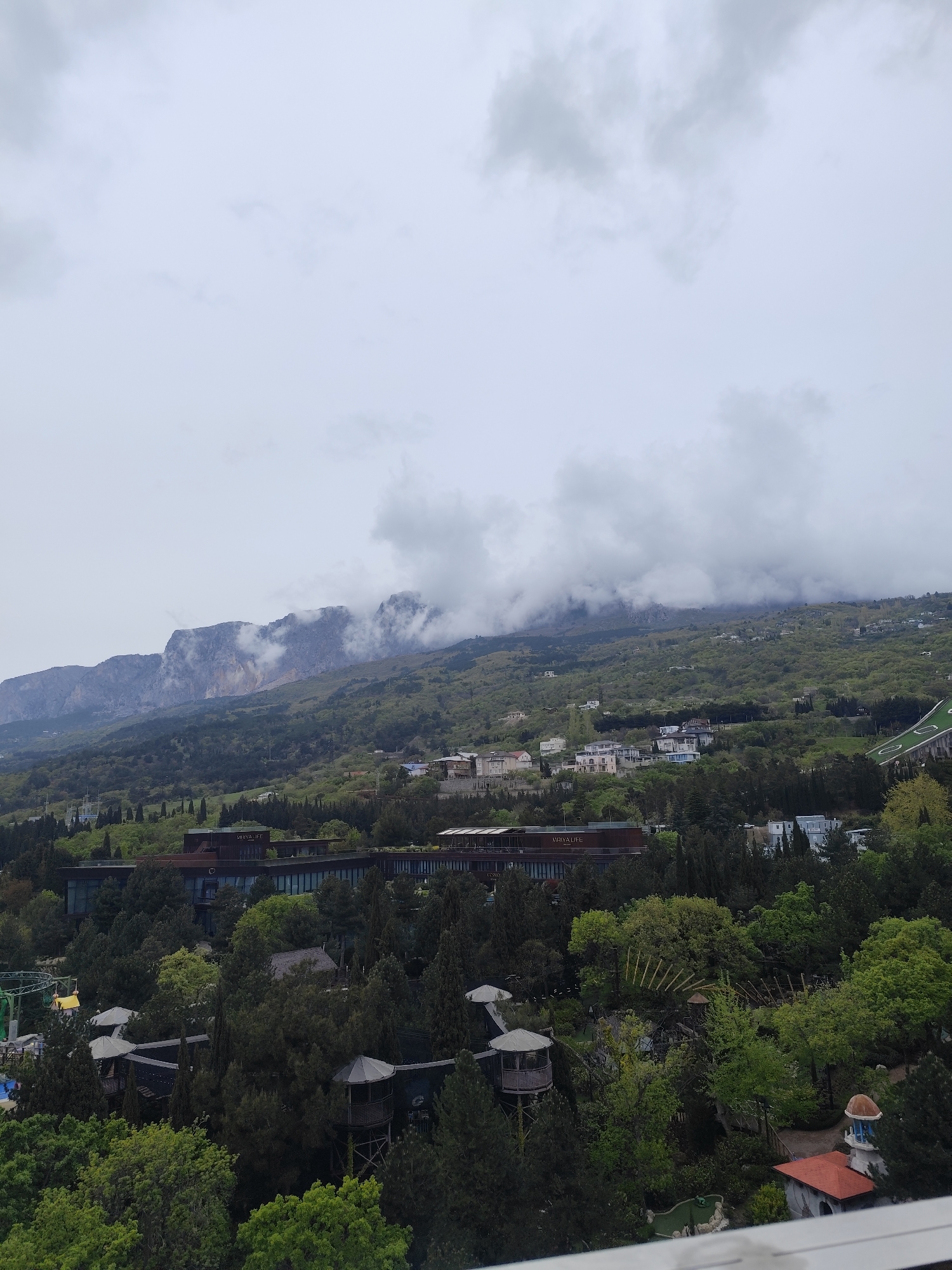 The image presents a wide view of a mountainous landscape under a heavily overcast sky. Rugged mountains dominate the background, their upper slopes and peaks partially obscured by dense, grey clouds, suggesting a misty or damp atmosphere. The hillsides leading down are covered in a rich tapestry of green vegetation, primarily dense forests and scattered trees.
In the middle ground, a large, modern building, likely a resort or hotel, is prominent among the trees. The text "MRIYA LIFE" is clearly visible on its facade, with "CONO" partially visible below it. Numerous other smaller residential buildings and houses with light-colored exteriors are scattered across the green slopes, blending into the environment.
The foreground and lower middle ground reveal elements of a recreational or amusement park, nestled within the dense foliage. These include several distinctive, pavilion-like structures that resemble gazebos or treehouses, a small area resembling a miniature golf course, and a large green slide or ramp on the right. To the far left, a yellow gondola or cabin suggests the presence of a cable car system. The sky is uniformly grey and cloudy, indicating an overcast day. The scene depicts a resort or leisure complex integrated into a verdant, mountainous coastal region, consistent with the provided location context of Симеизский поселковый совет, Ukraine. No specific human activity is visible.