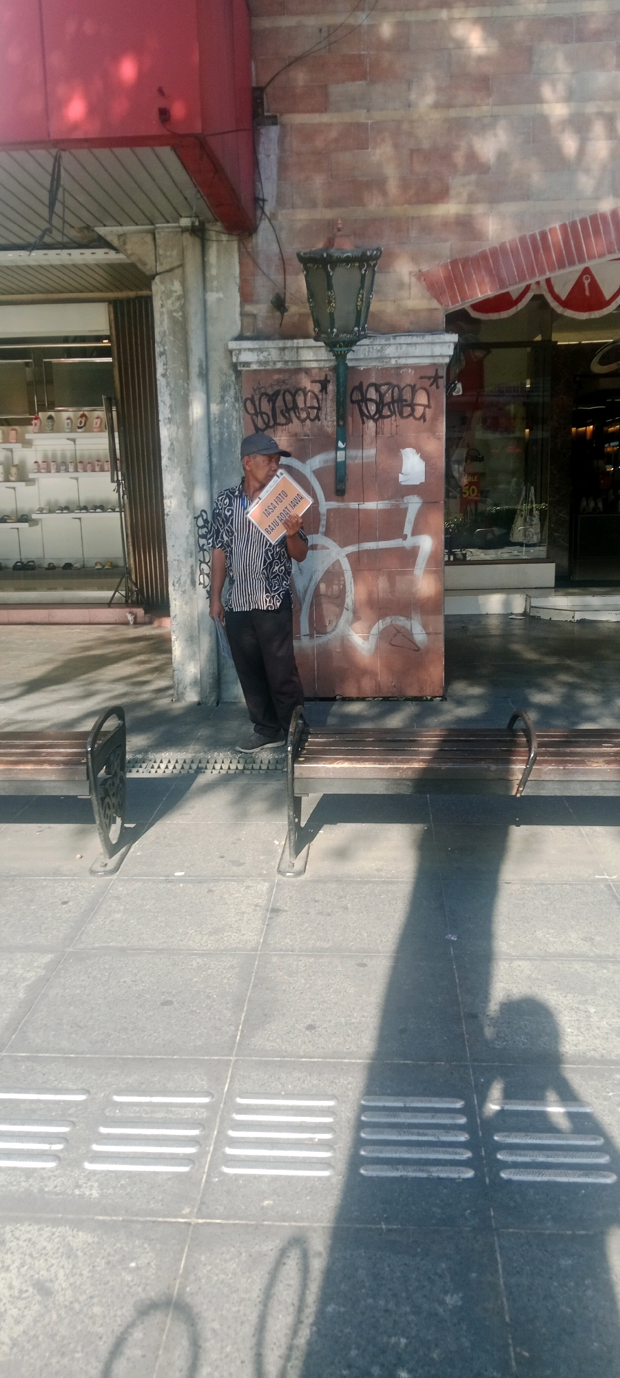 An elderly Indonesian man wearing a black and white striped shirt and dark pants stands on a paved sidewalk in front of a brick building with graffiti. He is wearing a dark baseball cap and holding a rectangular sign with orange borders that reads "JASA FOTO BAJU ADAT JAWA" in black lettering. He appears to be looking at the sign he is holding. To his left is the entrance to a store with a red awning. In front of the man are two wooden benches with black metal legs. The building behind him has a dark green lantern attached to the wall. The scene is illuminated by sunlight, casting shadows on the ground. This scene is located in Yogyakarta, Indonesia, likely on a street with commercial establishments.