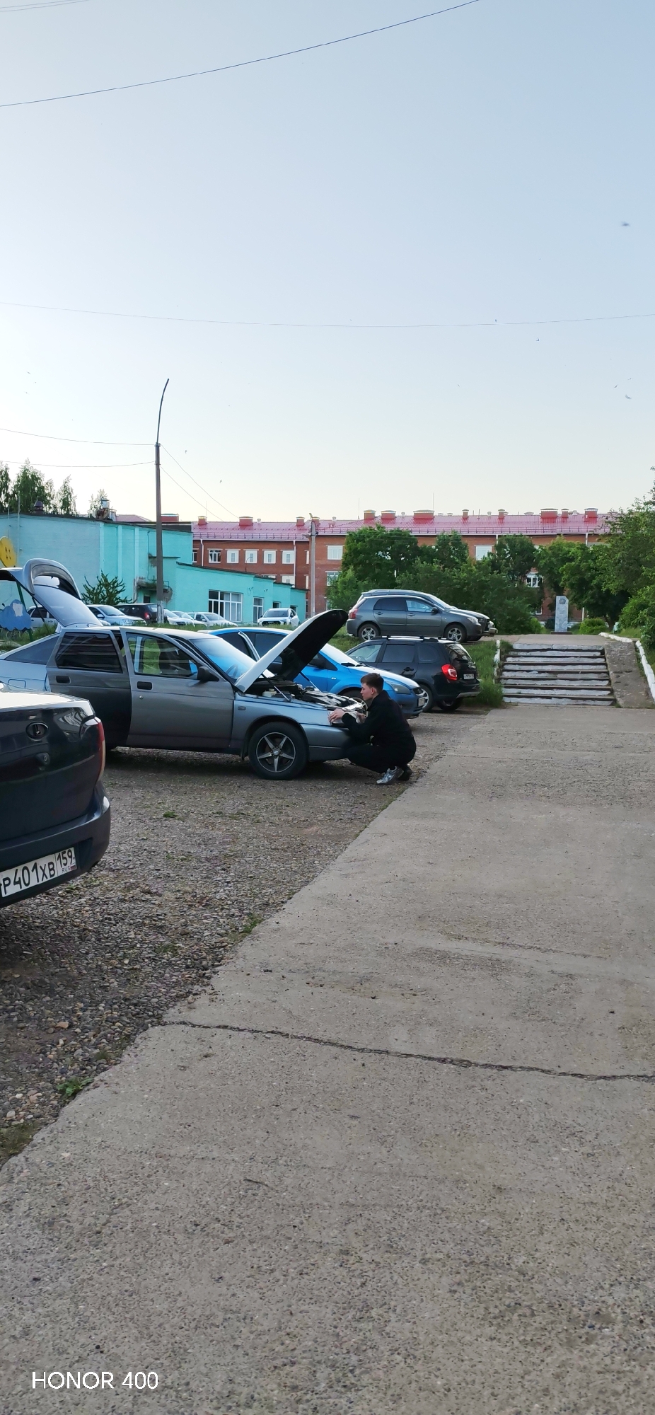 The photograph depicts an outdoor scene in what appears to be a parking or unpaved area in Noviy, Russia, under a clear, bright sky, likely during late afternoon or early evening.
The central subject is a grey four-door sedan, possibly a Lada model, with its hood propped open, suggesting a breakdown or maintenance activity. A person, appearing to be male and dressed in dark casual clothing with light sneakers, is crouched beside the front of this car, peering into the engine bay with an engaged expression, as if inspecting or working on the engine. The car's driver-side door is open, and the trunk is also slightly ajar.
Behind this grey sedan, another dark blue car also has its hood open. Other vehicles are parked around, including a dark grey SUV further back. In the immediate foreground, the rear of a black car is visible, bearing a Russian license plate that clearly reads "P401XB 159 RUS". The "159" region code corresponds to Perm Krai.
The ground is a mix of gravel and concrete, with a concrete pathway extending from the bottom right. In the background, there are multi-story buildings, including one with a turquoise facade and others constructed of red brick with matching red roofs, typical of Russian architecture. A set of concrete stairs with handrails leads uphill on the right side of the frame. A tall street light pole is visible on the left.
The scene conveys a sense of everyday life, possibly capturing an impromptu repair or check-up. The visible text "HONOR 400" is imprinted in the bottom left corner, indicating the camera or device used to take the picture.