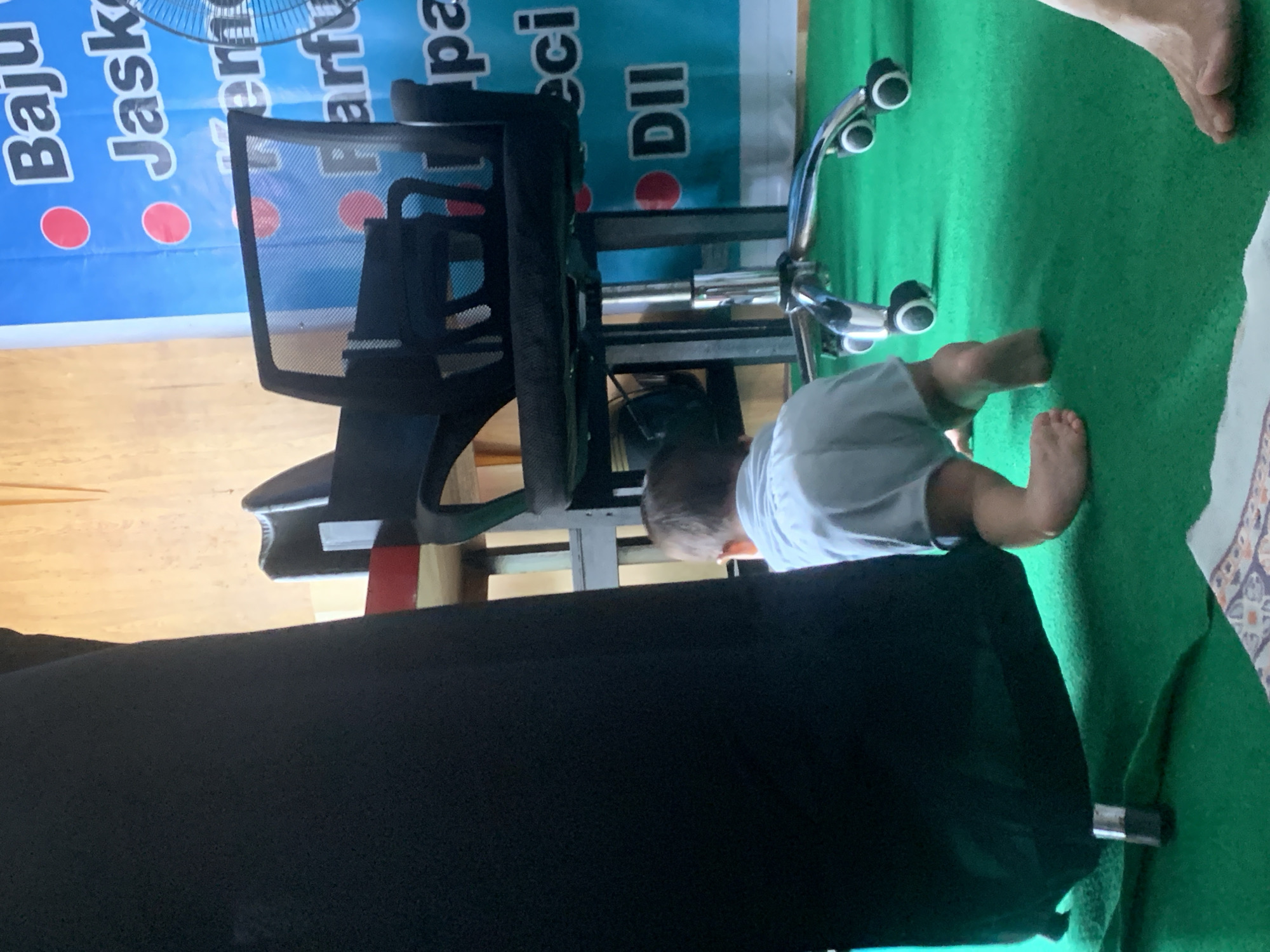 The image shows a child lying on their stomach on a green carpeted floor, playing near a black office chair with a metal base. The child is wearing a white t-shirt and light-colored shorts. Their legs and feet are visible, and they appear to be looking down at something.
Behind the child and the chair is a blue banner with text in white and red. The visible text on the banner includes "Baju Jaske", "erfect", "pa", "eci", and "DII". A fan is partially visible at the top left of the banner.
To the left of the banner, a wooden surface is visible, likely a table or floor. The overall setting appears to be indoors, possibly a room in a home or a community space. The lighting suggests it could be daytime. No specific weather conditions are evident. The child's activity is one of casual play or exploration.