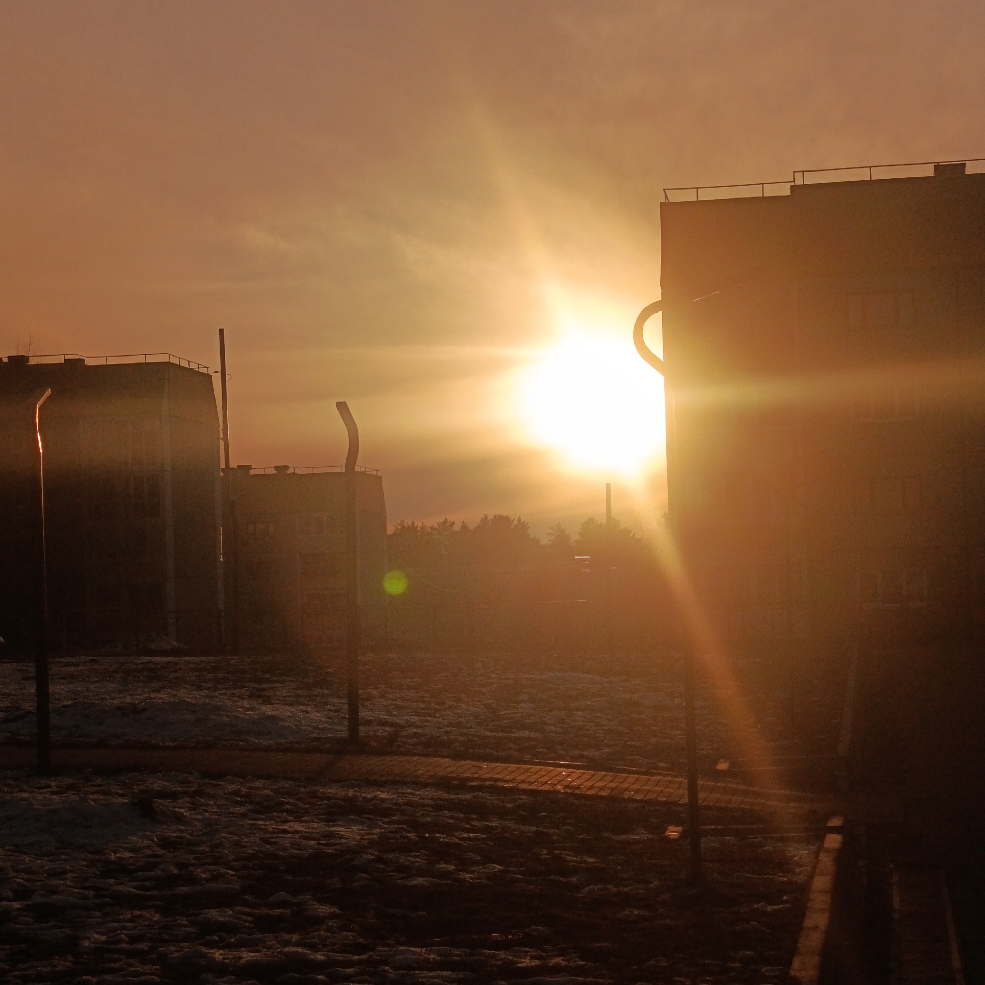 The image depicts a bright, possibly hazy sunset in Markovskii, Russia. Several buildings are visible, silhouetted against the light. In the foreground, there is ground covered in what appears to be a mix of snow and dirt. Several utility poles are present. The sun is positioned centrally in the image, creating a strong glare and long rays of light. The sky is orange.