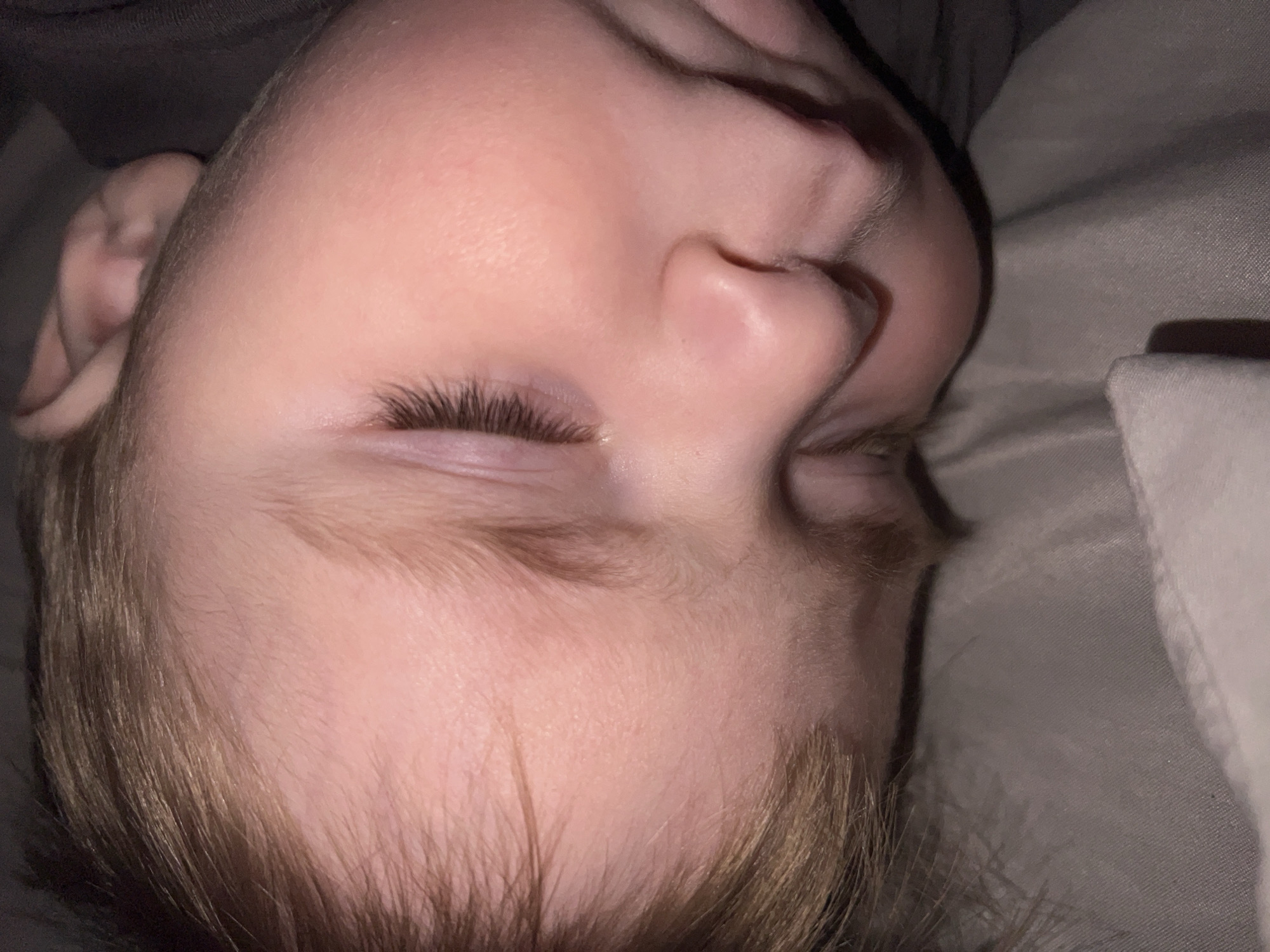 The image captures a close-up, overhead view of a young child sleeping or resting peacefully. The child lies on their side, with their head slightly tilted upwards, eyes gently closed. Their fair skin appears smooth, and light brown or blonde hair frames their face, visible on the top and side of their head. Long, dark eyelashes are prominent against their closed eyelids, with a subtle hint of redness visible on the lower lid of the closest eye. A small, upturned nose is visible in profile.
The child is partially covered by or lying on a dark-colored garment, possibly a shirt or blanket. They are resting on light grey or beige fabric, likely a bedsheet or blanket, suggesting an indoor setting, most probably a bedroom. The lighting is dim, indicating either nighttime or a softly lit room, contributing to a serene and tranquil atmosphere. No specific location cues or text are visible within the frame. The child's expression conveys a sense of calm and deep slumber.