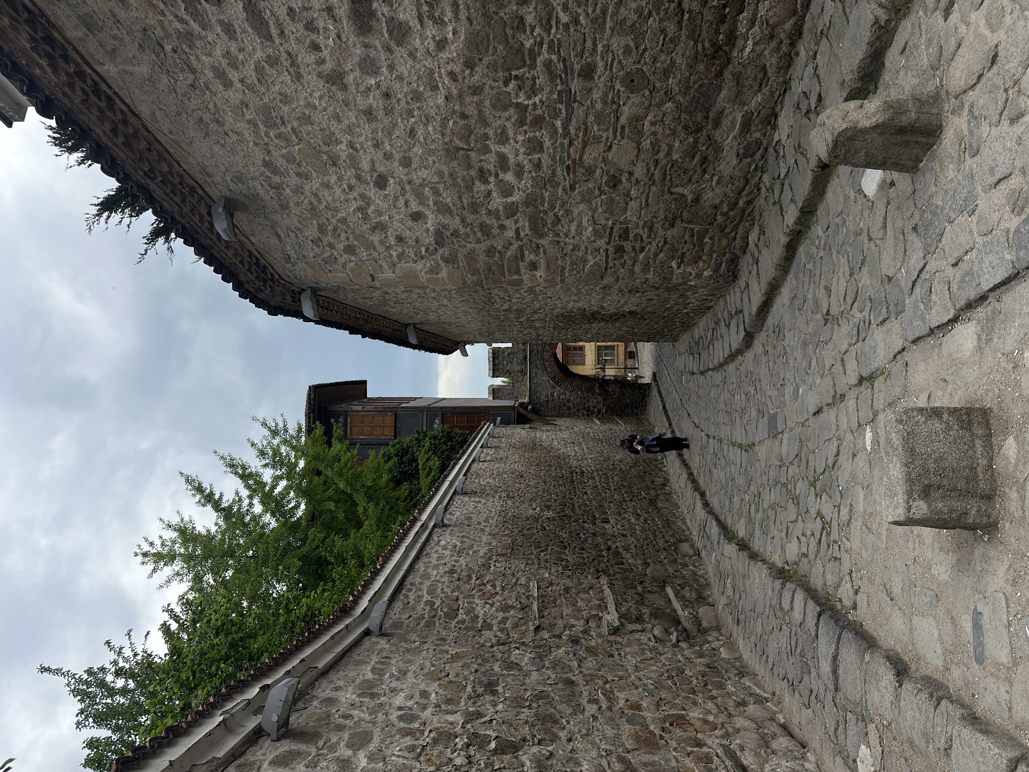 A narrow cobblestone street is framed by stone walls, indicative of a historic area, likely within the city of Plovdiv, Bulgaria. A person walks away down the street, between the tall walls, under a cloudy sky. Vegetation hangs over the wall on the left side of the image. Architectural details such as roof tiling and stone brick are visible.