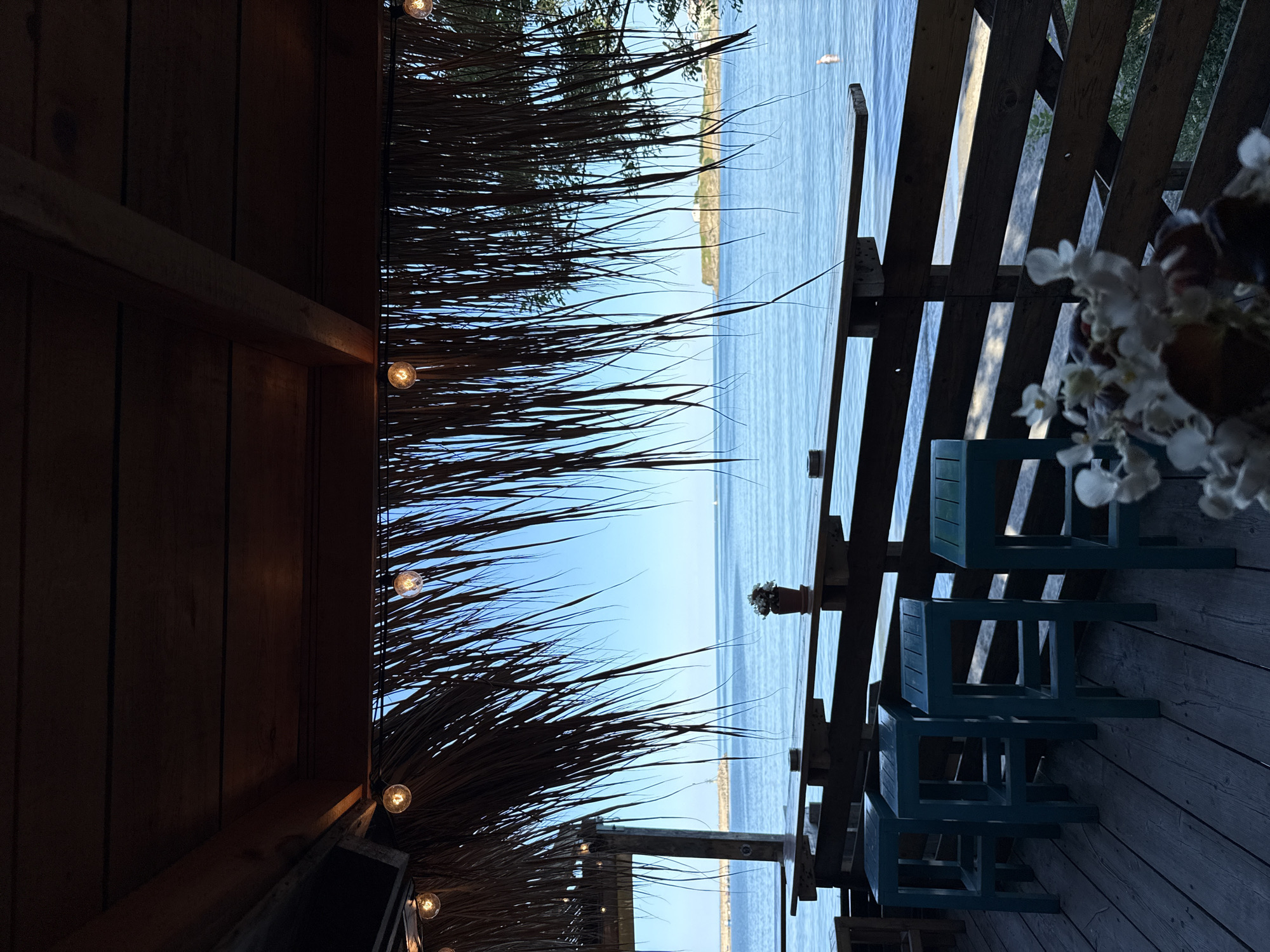The image shows a view of the sea from a covered outdoor seating area. The seating area has a thatched roof and is furnished with a long wooden bar and several teal-colored stools. The bar overlooks the sea, which is calm and blue. In the distance, a small island with a building on it can be seen. The scene appears to be a waterfront cafe or restaurant in Tsarevo, Bulgaria.
