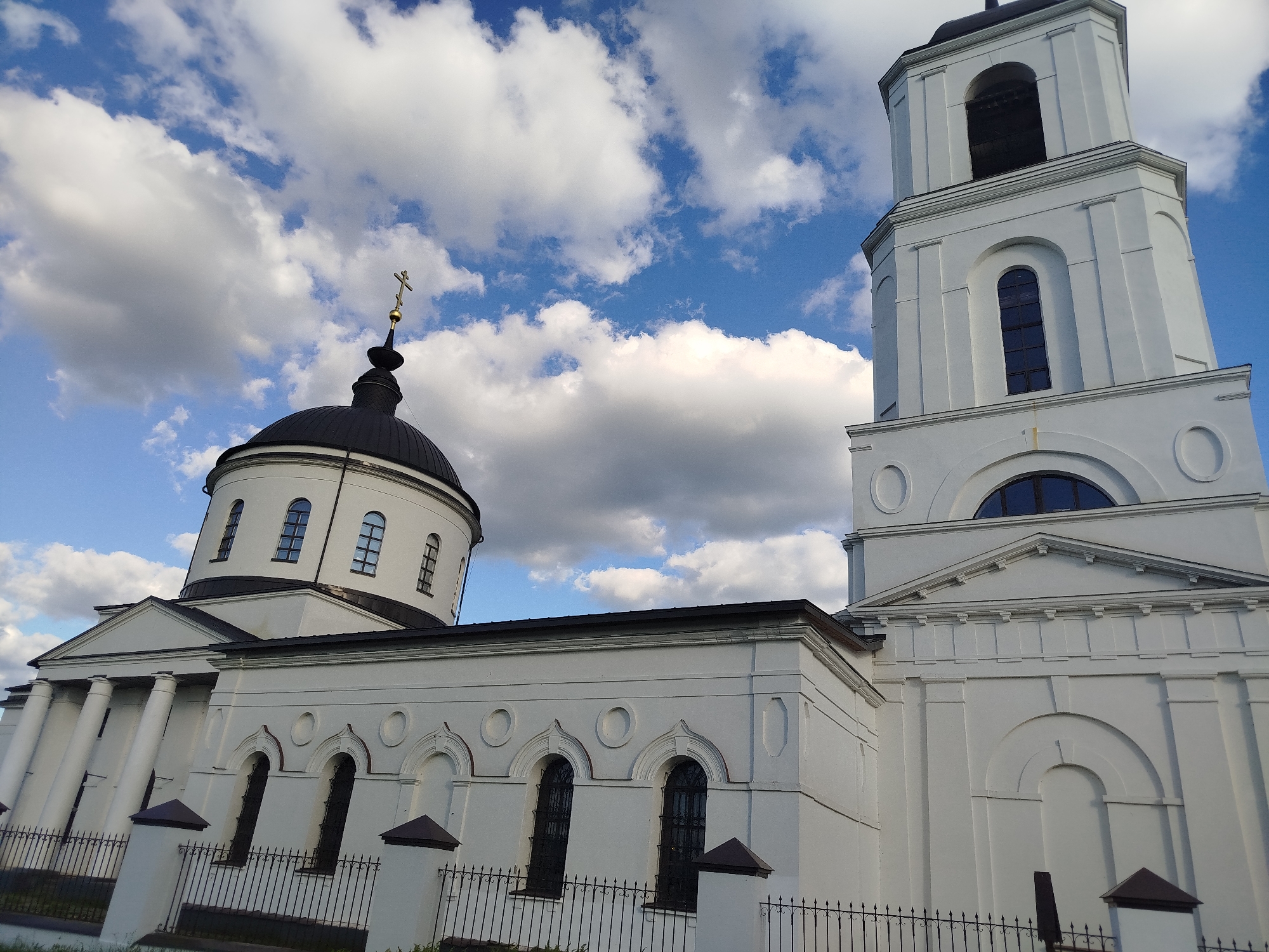 The image shows a low-angle view of the Church of the Intercession in Боровково, Russia, set against a sky with scattered white clouds and patches of blue. The church is predominantly white with a black dome topped with a golden cross. Architectural details include columns, arched windows, and a tall, rectangular bell tower to the right. A metal fence runs along the base of the building.
