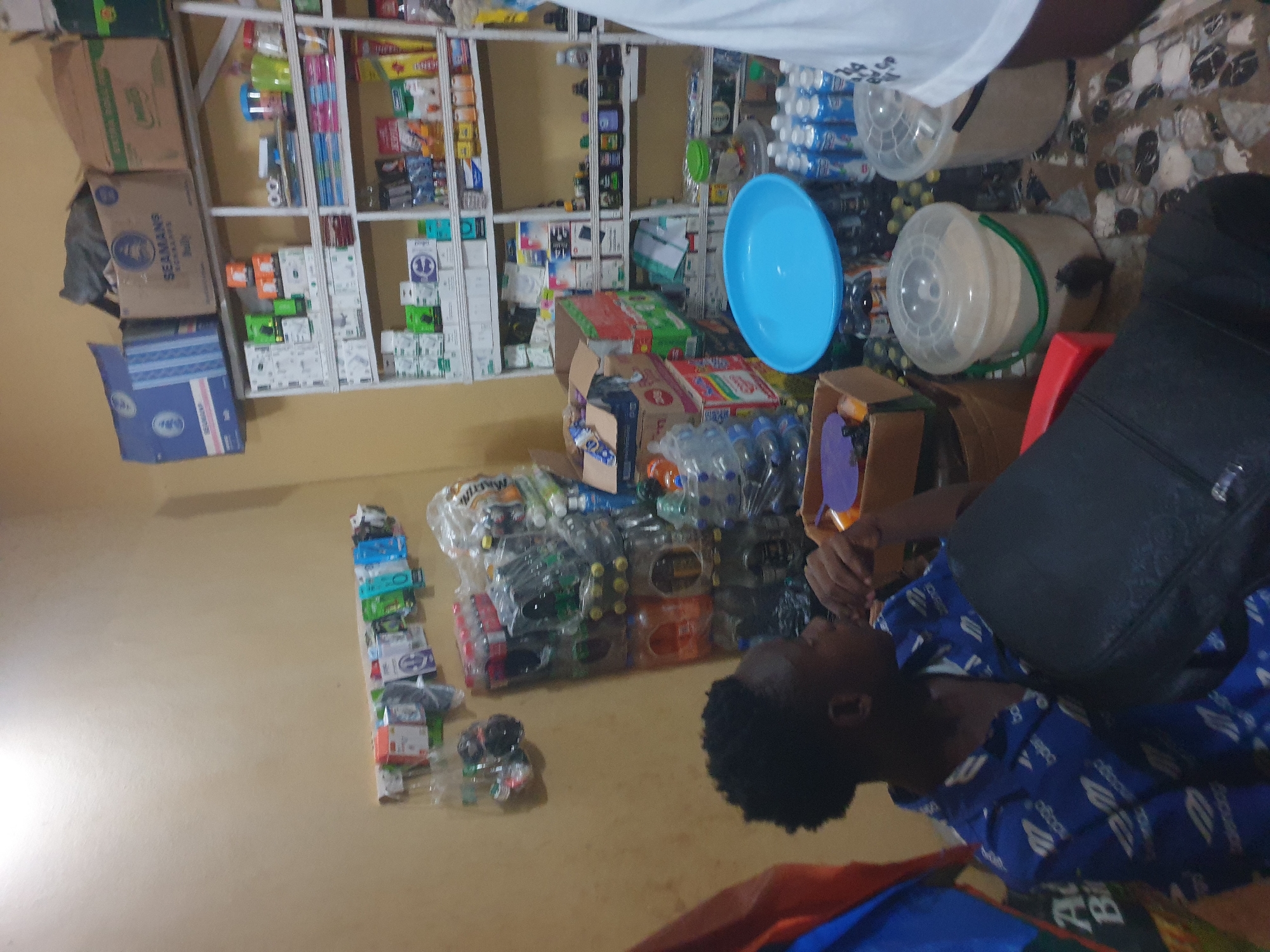 The image depicts an interior scene within a small shop in Anilelerin, Nigeria. A young Black male, wearing a blue patterned shirt, is seated in the foreground, partially obscured by a black bag. His right hand is near his mouth, and he appears to be looking to the right, away from the camera. The shop is densely stocked with various items, including bottled beverages, packaged goods, and what appear to be cleaning supplies or toiletries, organized on shelves and in stacks. A blue plastic basin is visible on a shelf, along with several large plastic buckets filled with granular material. The walls are painted a light color, and the floor appears to be tiled with small, irregularly shaped stones. The overall impression is that of a busy, well-stocked convenience store or local market stall.