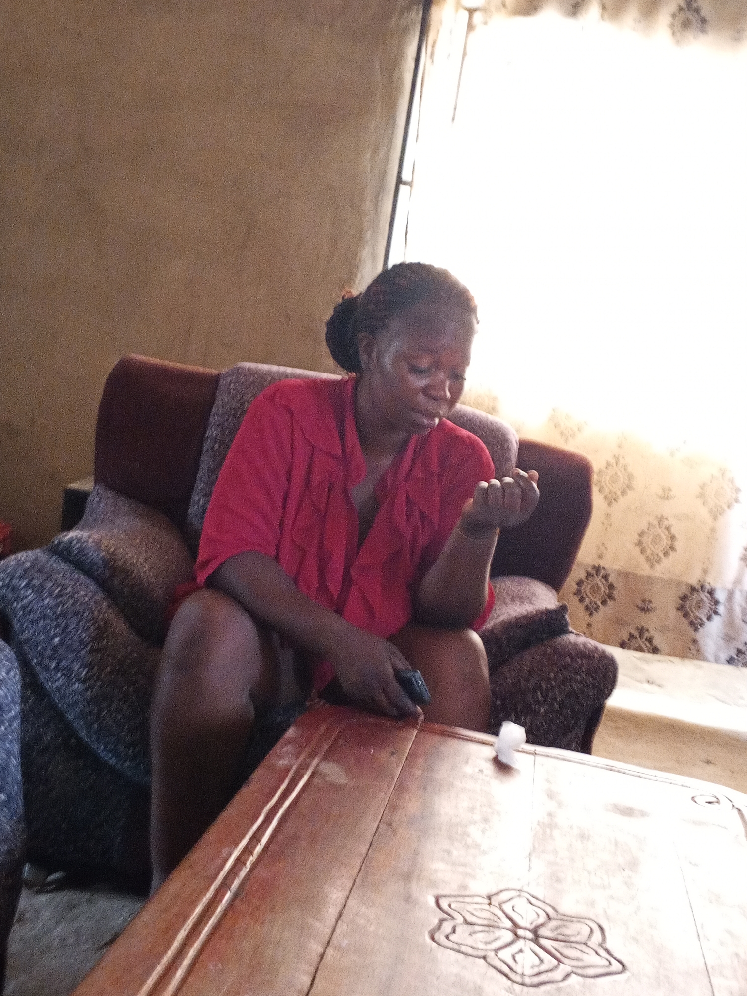 The image captures an indoor scene, likely a living room, featuring a woman seated on a sofa. The woman, an adult with dark skin and dark hair pulled back, is wearing a bright red, short-sleeved top with ruffles. She is seated in a maroon and patterned grey armchair, leaning slightly forward with her gaze directed downwards. In her left hand, she holds a small, dark object, possibly a phone or remote control, while her right hand is open and resting near it. Her expression appears focused or pensive.
Abundant natural light streams in from a window positioned to the right of the woman, causing an overexposed area and creating some backlighting. Light-colored curtains adorned with a subtle floral or geometric pattern are visible near the window. The room's walls are a textured, light brown or tan color.
In the foreground, a prominent wooden table with a light brown or reddish-brown hue is visible. It features a distinct carved floral design, resembling a lotus, in its bottom right corner. A small, crumpled white object, possibly a piece of tissue or cotton, rests on the table's surface. The setting suggests a casual, everyday moment. There is no visible text in the image.