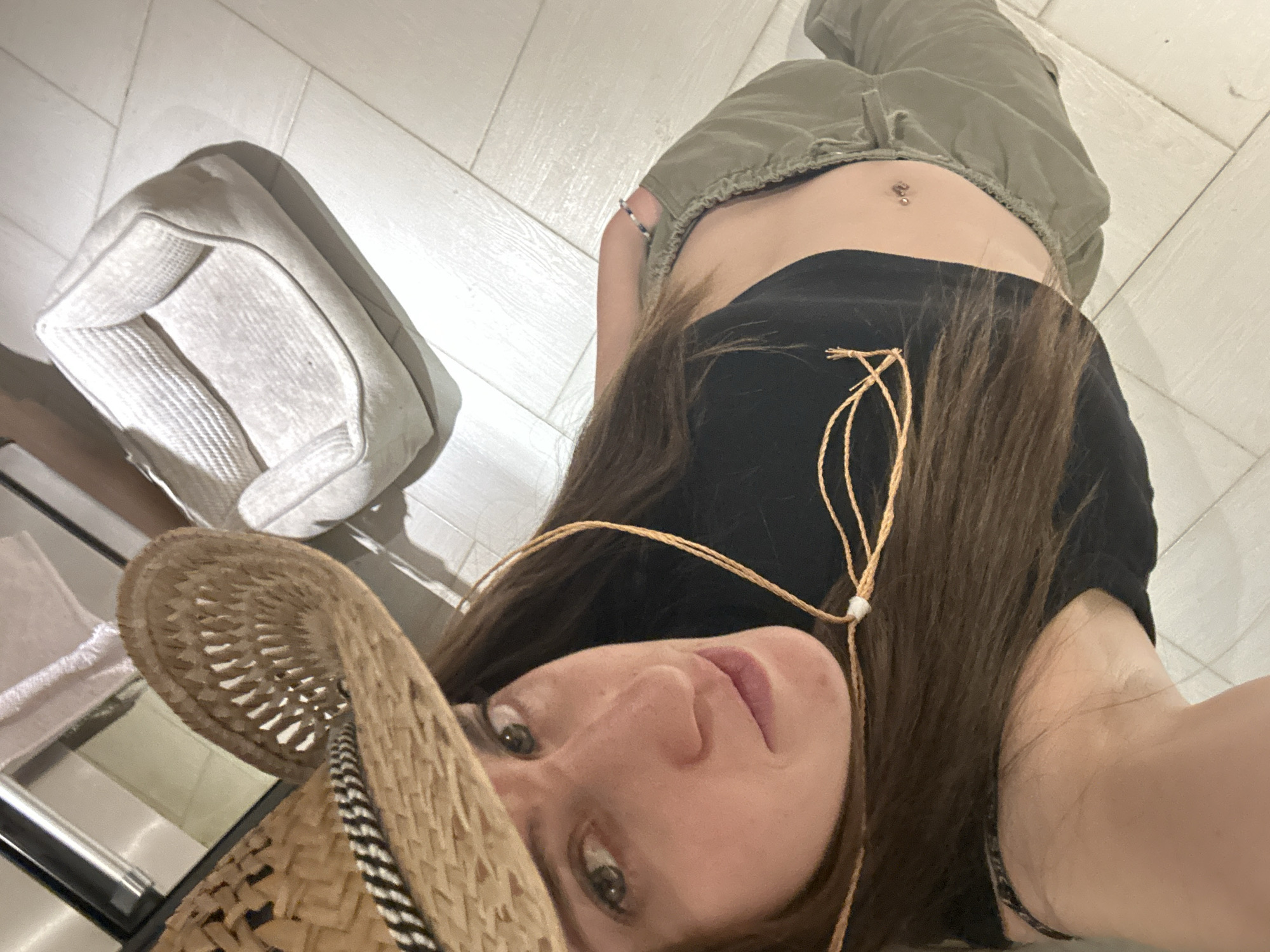 A woman is depicted in a selfie-style image. She is wearing a black top, green pants, and a straw cowboy hat. Her midriff is visible, and she has a navel piercing. She is looking directly into the camera. A light-colored upholstered chair is visible in the background. The floor appears to be tiled. The location appears to be indoors. There is no indication that the location is in Mexico City, Mexico.