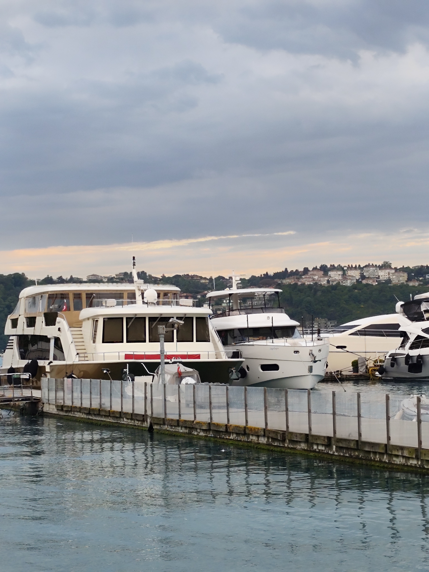 The image depicts a marina scene in Sarıyer, Turkey, characterized by several yachts docked along a weathered pier. The sky is overcast with a gradient of light to dark gray clouds, suggesting an early morning or late afternoon.
The boats are of various sizes and designs, ranging from a multi-tiered yacht with a tan exterior to sleek, modern yachts with white and gray hulls. The pier, constructed from wood and transparent plastic panels, shows signs of aging with visible wear and moss growth. The water in the marina reflects the colors of the sky and the pier, creating a rippled effect. In the background, a distant cityscape is visible, with buildings situated on a green hillside, typical of the Sarıyer landscape. There is a single person visible in the image on the edge of the pier on the left side.