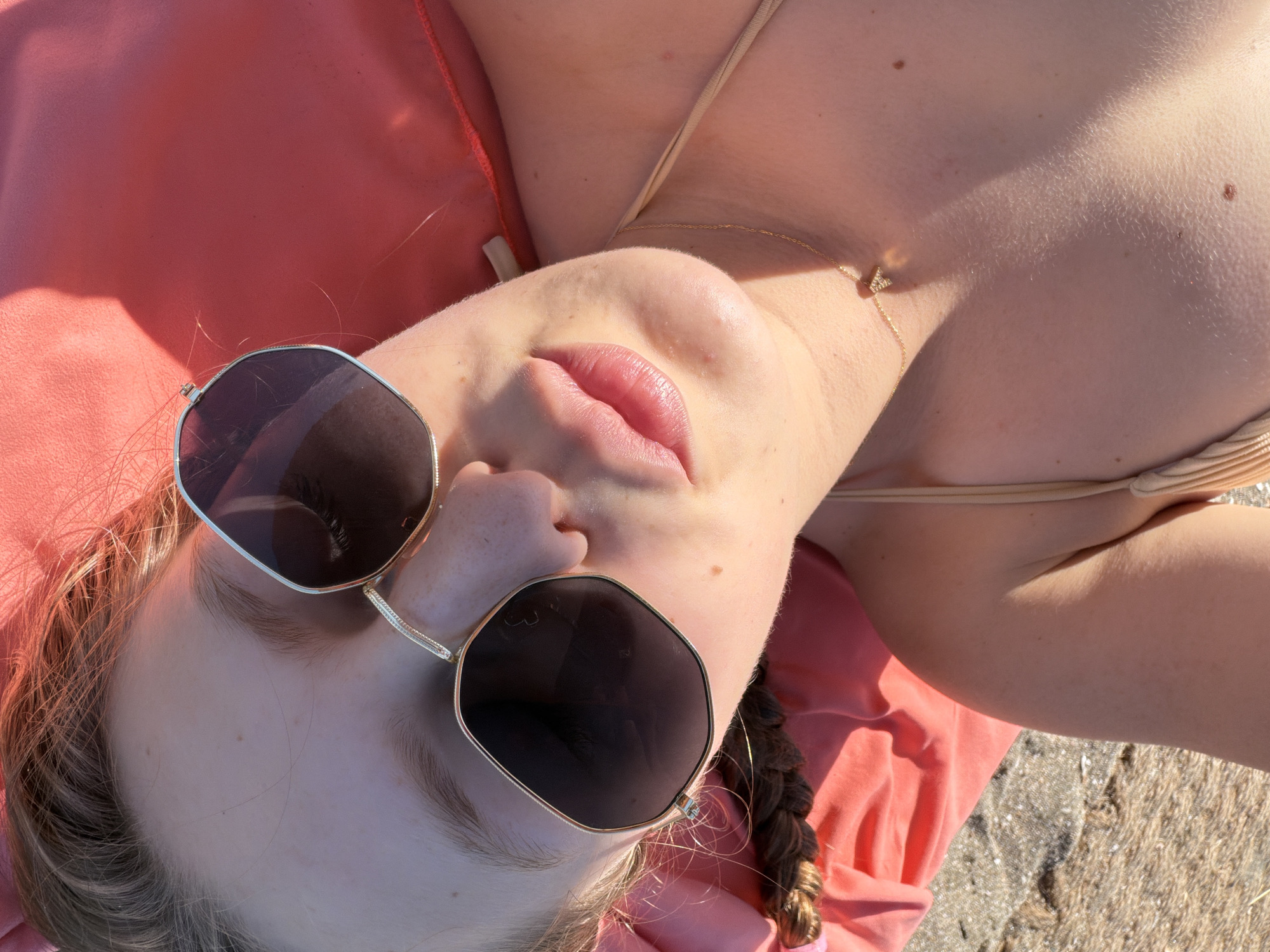 The image shows a close-up, overhead view of a young woman lying on a pink surface, possibly a towel or cushion, on a sandy beach. She is wearing large, hexagonal, gold-rimmed sunglasses with dark lenses, and a beige bikini top. A delicate gold necklace with a small pendant is visible on her collarbone. Her hair, in a braid, is partially visible to the left of the frame. The lighting suggests it is a sunny day, with shadows cast across her face and neck. The background is blurred, showing sand and the pink surface. The overall impression is of a relaxed, sunbathing scene. The location is specified as a beach in Burgas, Bulgaria.