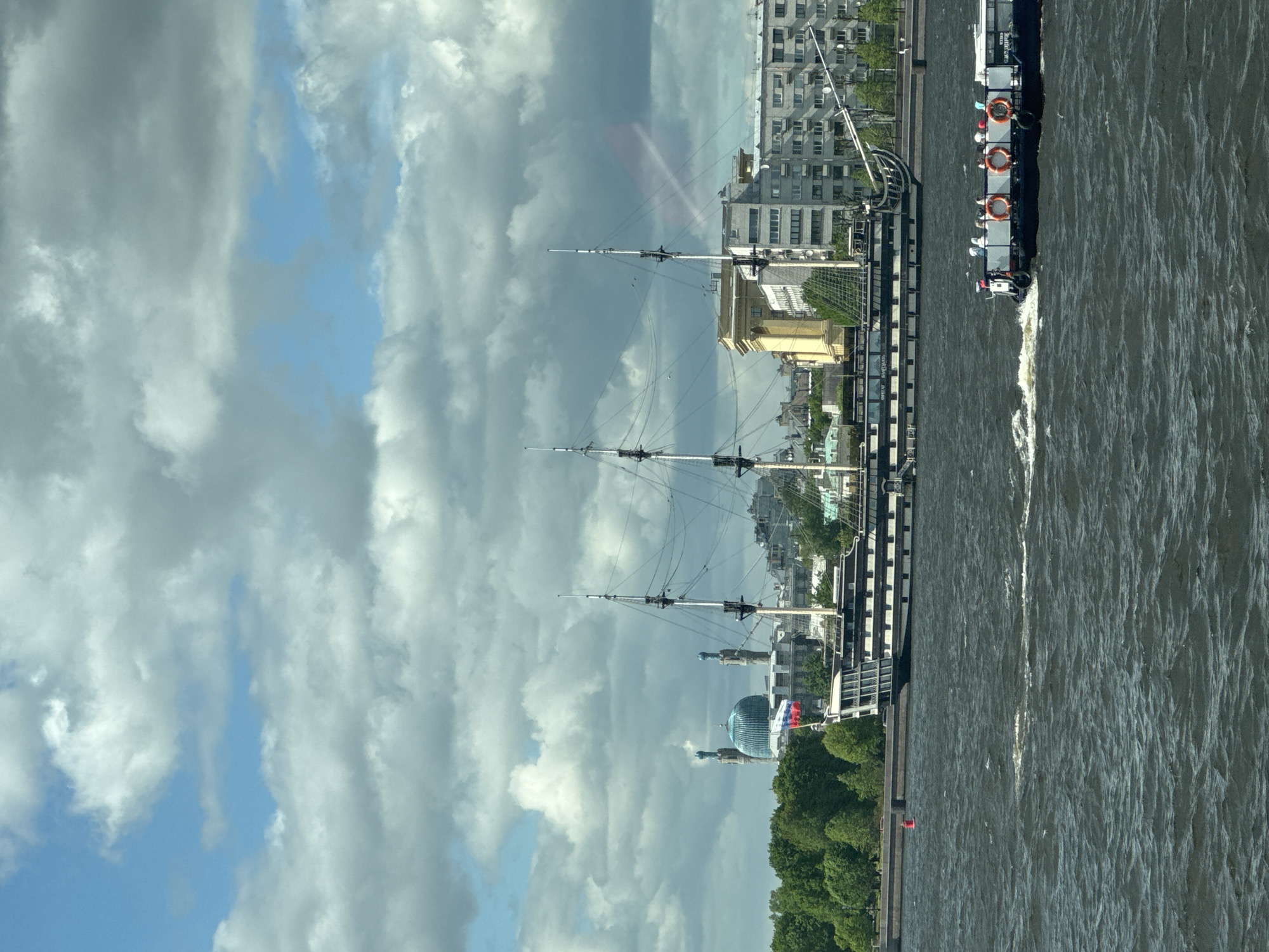 The image shows a view in Saint Petersburg, Russia, along the Neva River. A replica of the sailing ship "Poltava" is docked along the embankment. A tour boat is seen pulling away from the dock on the right. In the background, the dome of the Planetarium can be seen among the trees. The sky is cloudy.