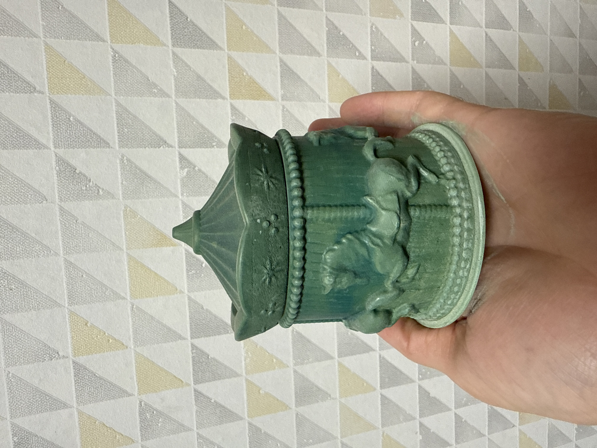 In an indoor setting with a geometric patterned wallpaper, a person's hand holds a small, dark teal carousel-shaped decorative box. The box features intricate detailing of horses and ornate trim. The wallpaper has a repeating pattern of triangles in shades of white, light gray, and pale yellow.