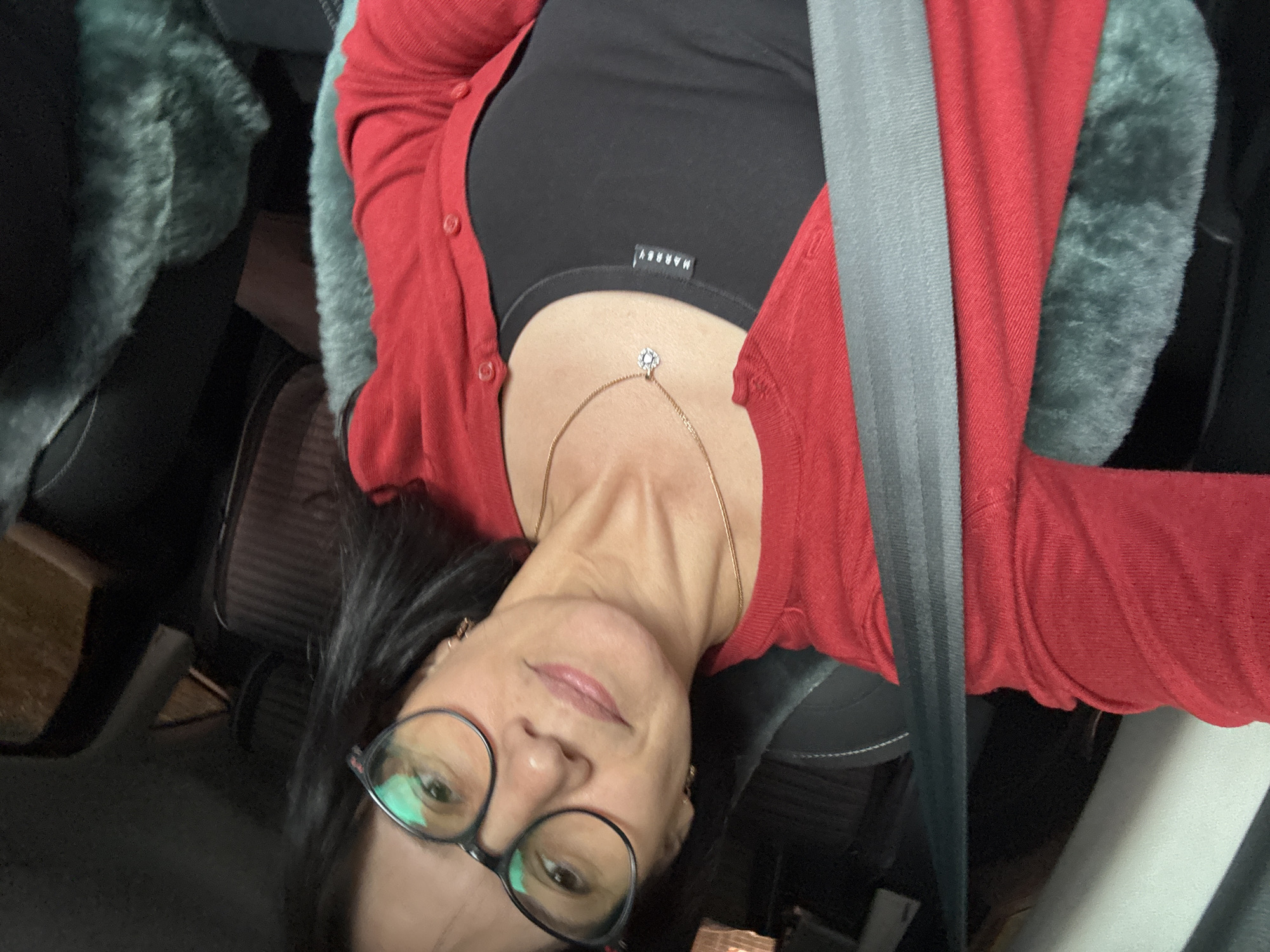 An image, rotated 90 degrees counter-clockwise, depicts a woman seated in what appears to be the interior of a car. Her head is positioned on the left side of the frame, and she is looking directly towards the camera with a neutral or slightly smiling expression.
The woman has dark hair and wears dark-framed glasses. She is adorned with a delicate gold chain necklace featuring a small, sparkly, flower-shaped pendant, and small earrings are visible. Her attire consists of a red button-up cardigan over a black tank top or camisole. A white label on the black top is visible, bearing the upside-down text "MARBEY".
A grey car seatbelt diagonally crosses her torso, from her right shoulder to her left hip. In the background, behind her, a textured, fluffy grey or teal material is noticeable, possibly a car seat cover or a blanket. Other parts of the car's interior, including dark upholstery and shadowed areas, are visible. The lighting suggests an indoor setting or nighttime. No specific location cues for Chaikovsky, Russia, are visible in the image itself, but this is the provided context.