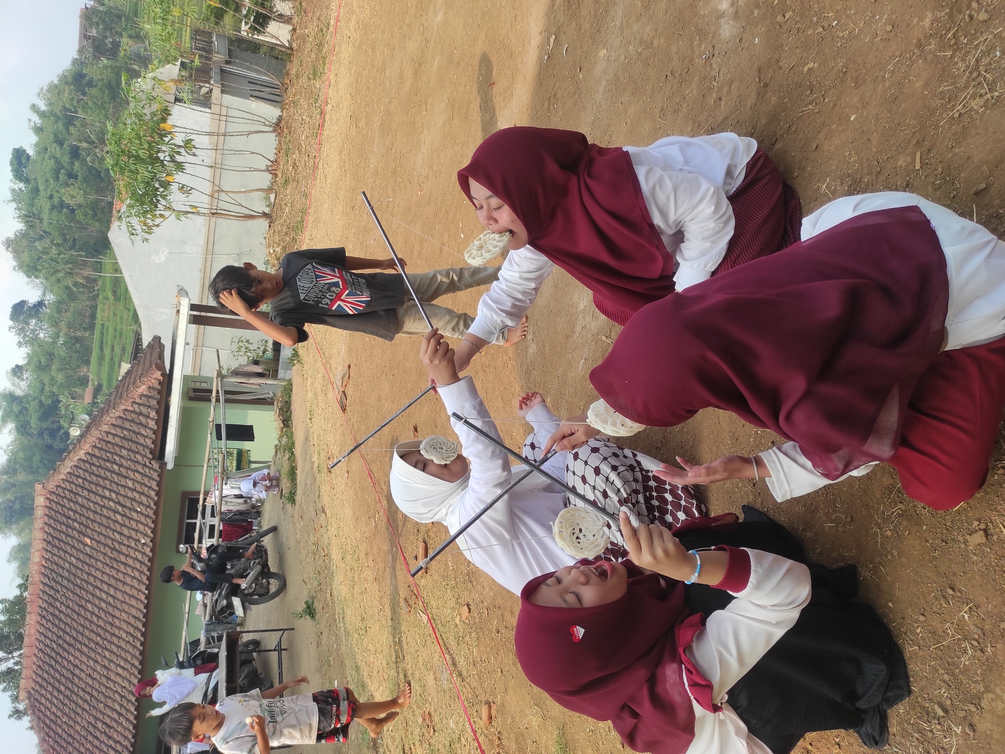 In Sumedang, Indonesia, four young girls are participating in a cracker-eating contest. They are seated on the ground, wearing hijabs and traditional Indonesian clothing. Each girl holds a cracker suspended by a string from a stick, attempting to eat it without using their hands. The contest appears to be taking place outdoors in a dirt-covered area, with a house and greenery in the background. Other children and adults are present, observing the activity. The scene suggests a community gathering or a festive occasion.