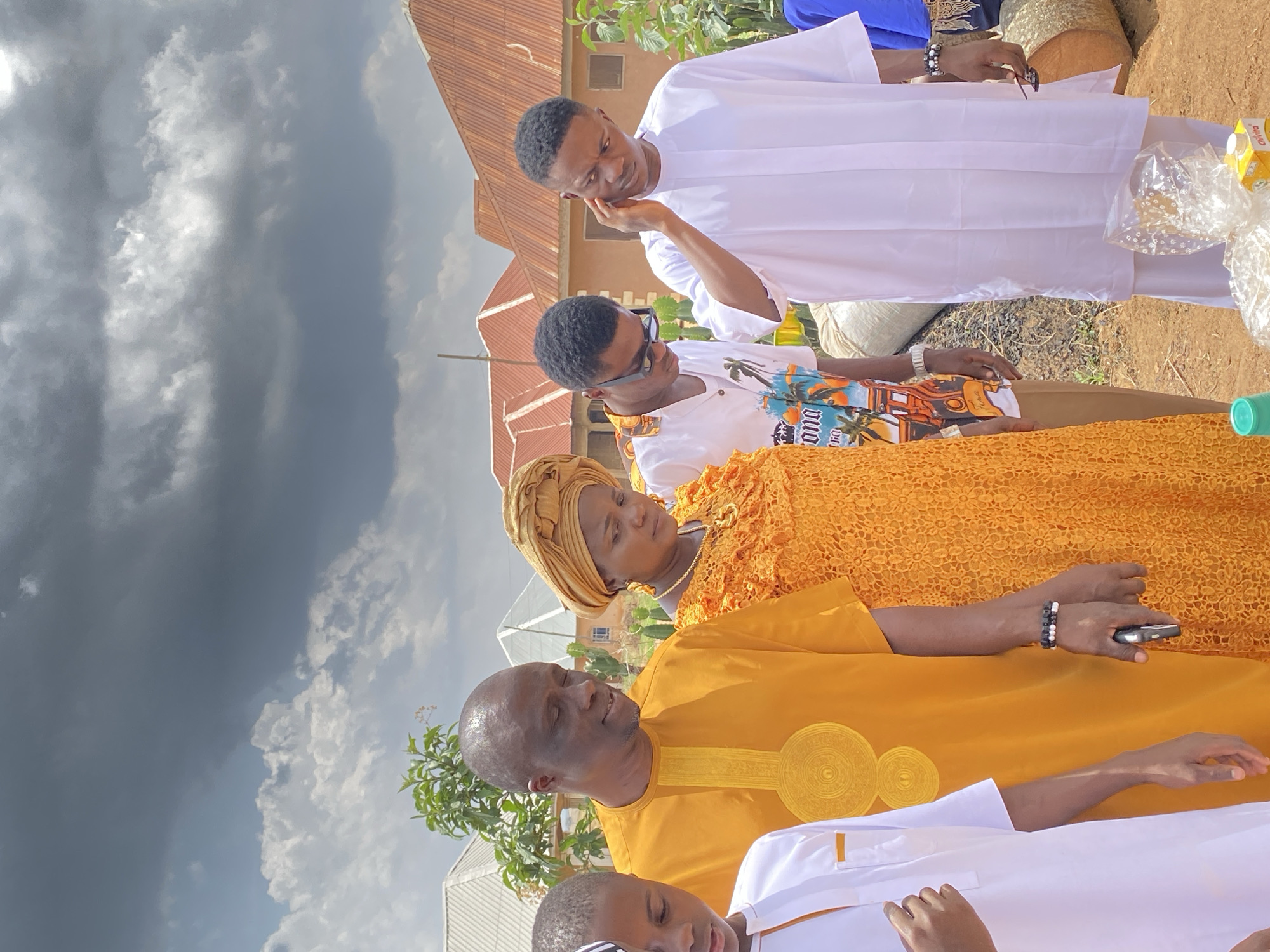 A group of people are gathered outdoors in Kuru Karama, Nigeria. The foreground shows part of a child’s head with a cap, then a man in a yellow traditional garment with decorative circles, and next to him, a woman in a yellow lace dress and head wrap. Behind them, another man is wearing a white short-sleeved shirt with a colorful print. A man in a white traditional outfit stands at the back. The people appear to be interacting, as they are close to each other. Some are looking towards the center of the group. The background includes houses with corrugated metal roofs, and the sky is cloudy and grey.