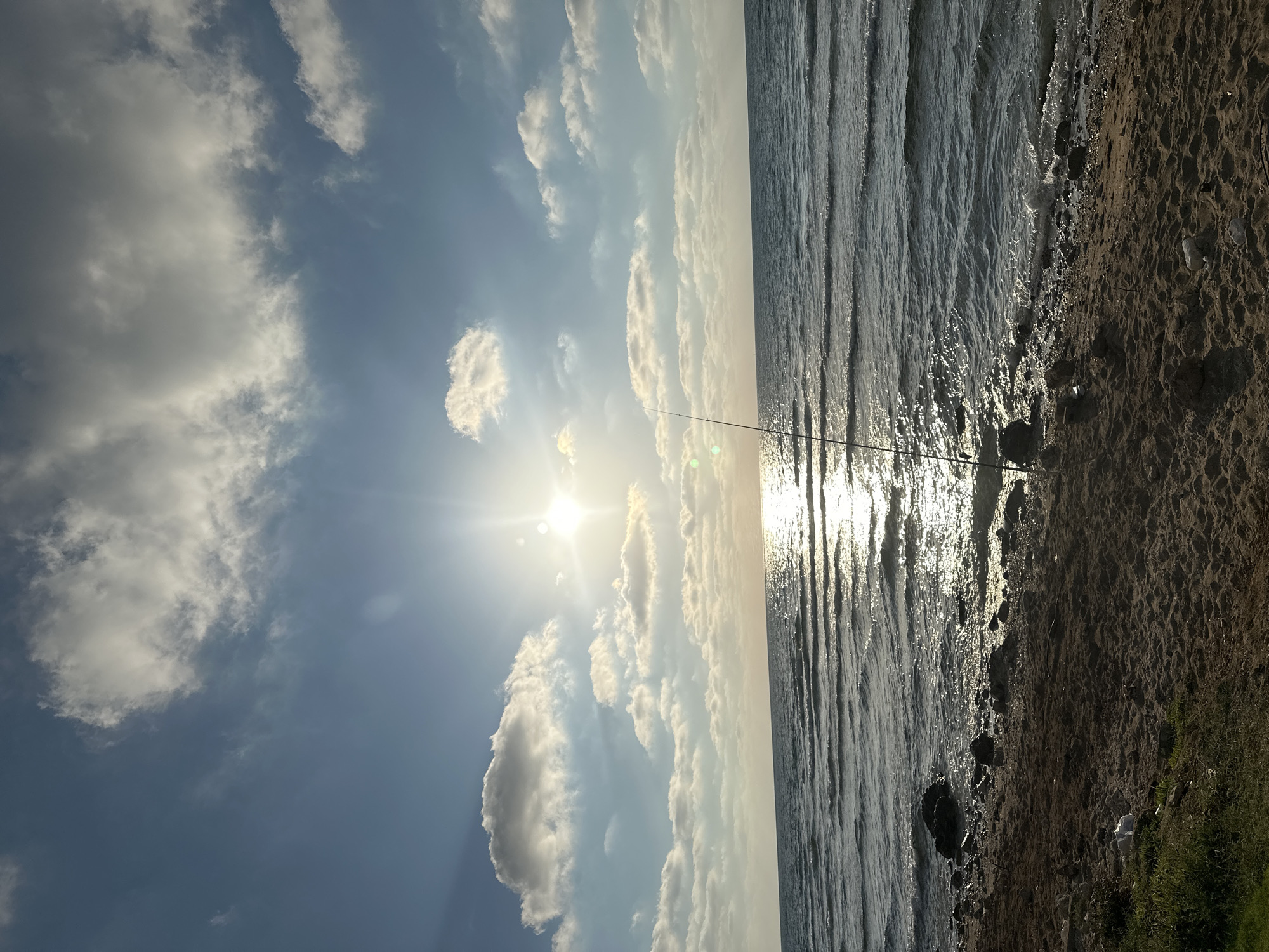 The image depicts a tranquil beach scene with the sun shining brightly in a partly cloudy sky. The sun's rays create a shimmering reflection on the gentle waves of the sea. A fishing rod stands upright on the sandy shore, suggesting someone is fishing. The beach is made up of sand and scattered rocks, with some patches of green grass on the left side. The sea is calm, with small waves breaking near the shore. The overall impression is one of peaceful solitude. The specific location in Haifa, Israel, is not discernible from the image itself.