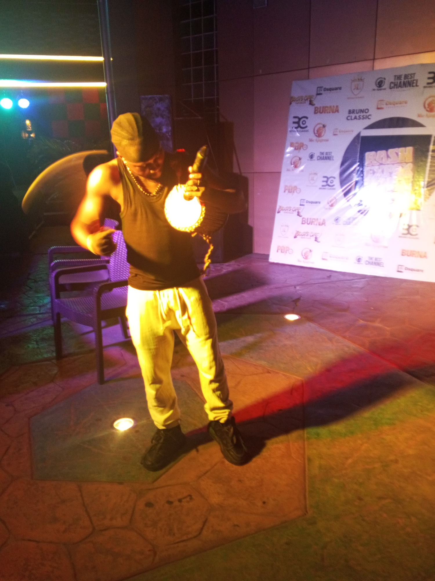 A man is standing on a stage in a room. He is holding a circular, illuminated object in his right hand. He is wearing a black tank top, gold chain, white pants, black shoes, and a black cap. Behind him is a chair and a sign with various logos and the words "BURNA", "BRUNO CLASSIC", and "THE BEST CHANNEL." The room is illuminated with colored lights. The location is in Lokoja, Nigeria.