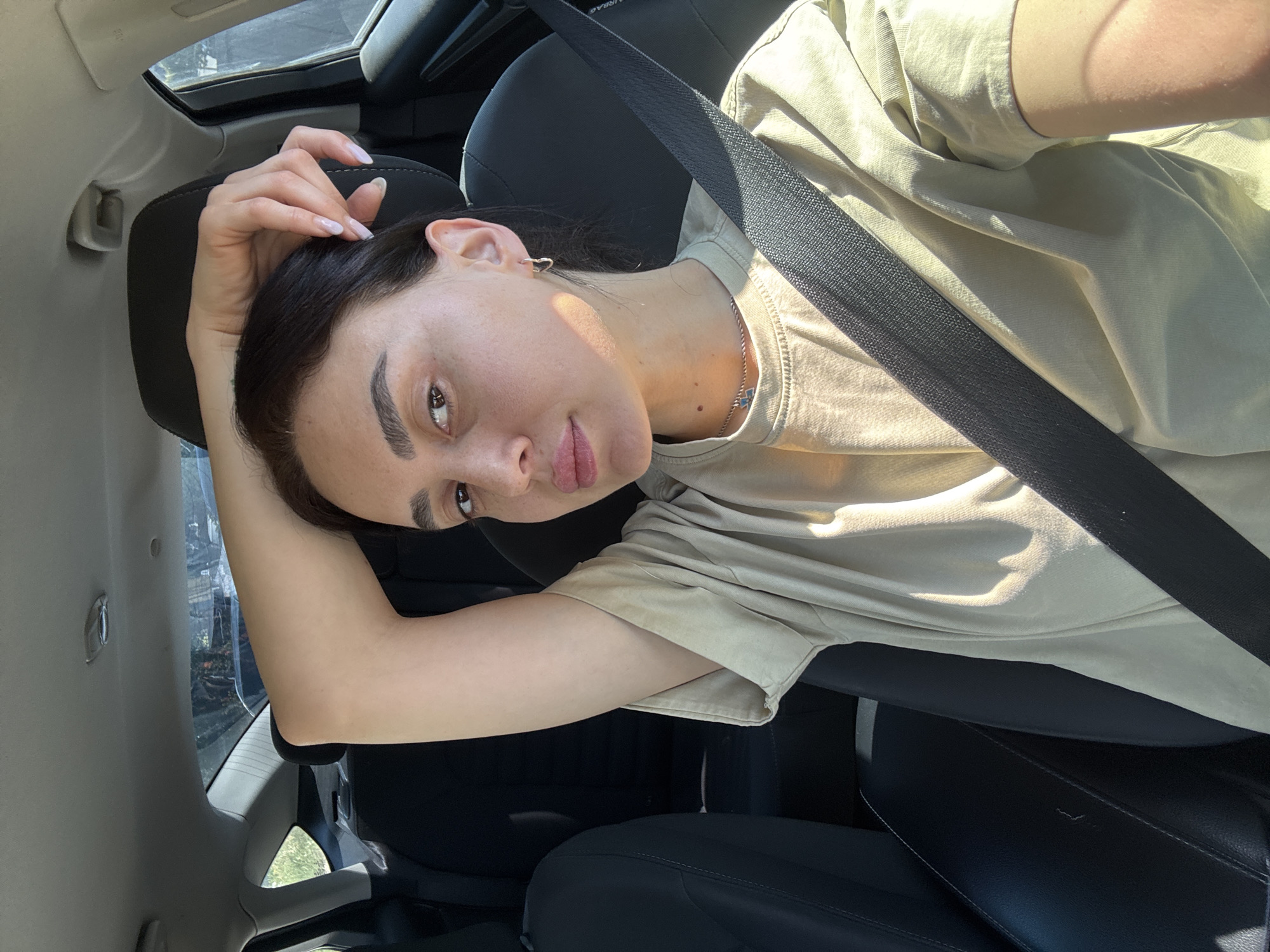 A young woman with dark hair is inside a car, wearing a tan short-sleeved shirt and a seatbelt, and looking towards the camera. Her right hand is above her head, resting near the car's headrest. A silver chain is visible on her neck. The car has black leather seats. The background is blurred, showing the interior of the car. The exact location in Tbilisi, Georgia, cannot be determined from the image.