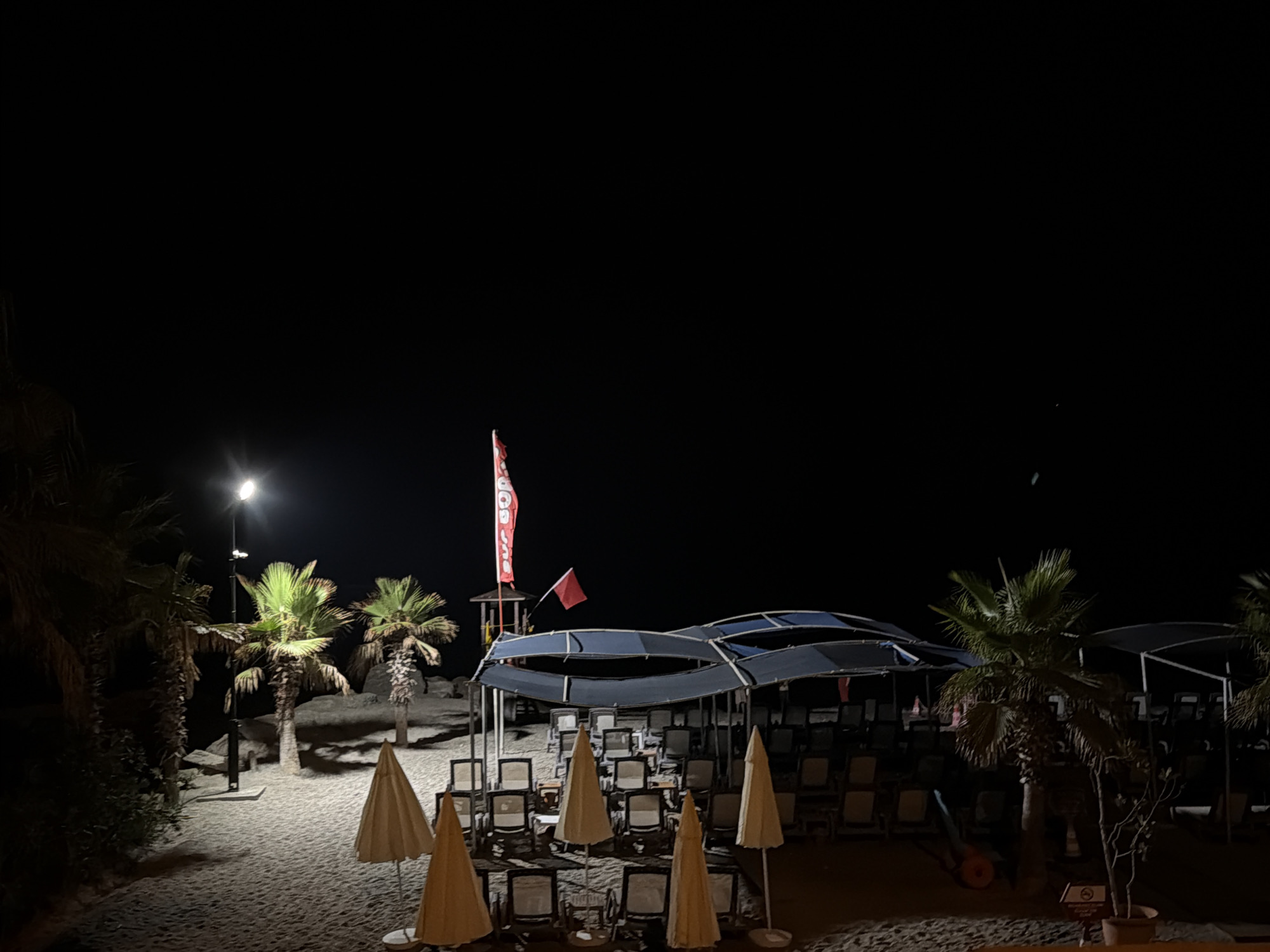 This is a nighttime photograph of a beach in Alanya, Turkey. The scene is dimly lit by a streetlamp in the upper left portion of the image, casting light on several palm trees and a sandy beach. In the background, a lifeguard tower with a red flag and a smaller red flag are visible. In front of the tower is a covered area with rows of empty lounge chairs, suggesting it is a resort or public beach that has closed for the night. Several closed beach umbrellas are also scattered across the sand. The sky is completely dark, indicating it is late at night. The overall impression is one of stillness and quiet after the day's activities have ended.