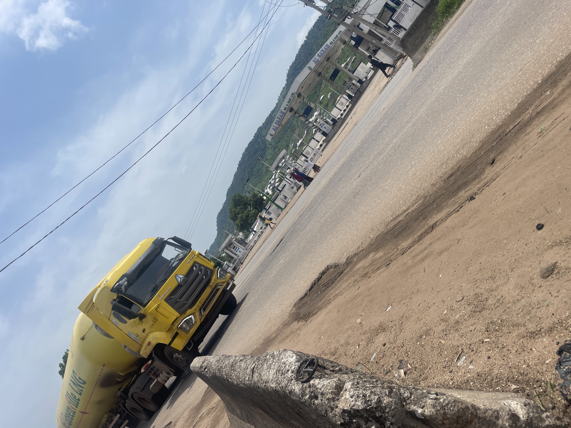 The image depicts a street scene in Lokoja, Nigeria. A bright yellow LNG (Liquefied Natural Gas) tanker truck is parked on the side of a road. The truck has "Greenville LNG" written in green on its side. In the background, a gas station with multiple pumps is visible, with the name "AL RABAH OIL & GAS LTD." displayed prominently on its signage. Beyond the gas station, a lush, green hillside rises, suggesting a somewhat mountainous or hilly terrain. The sky is partly cloudy with bright sunlight, indicating it is daytime, likely late morning or early afternoon.
The road appears to be a main thoroughfare, with tire marks and some wear visible on its surface, including a noticeable pothole. To the left of the road, there are some buildings and what seems to be a concrete barrier in the foreground. A few people are visible near the gas station and on the road, one person is walking away from the camera, and others are near the gas pumps. The overall impression is a busy, functional area where transportation and commerce are taking place under a clear, sunny sky. The presence of the LNG truck and gas station points to the importance of fuel distribution in the area.