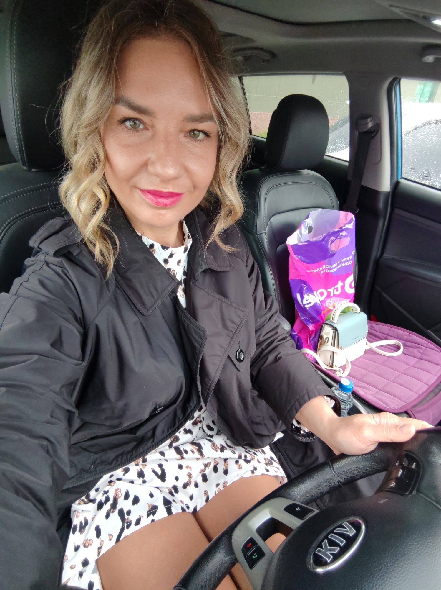 A woman with blonde curly hair, wearing a black jacket and a white dress with black and brown spots, is sitting in the driver's seat of a Kia car. She is holding the steering wheel. In the passenger seat, there is a purple seat cover, a purple and pink shopping bag with Cyrillic text, a small light blue and white handbag, and a bottle of water. The car's interior is black leather. It is raining outside, and droplets can be seen on the car windows. The photograph does not contain enough information to determine the exact location in the city of Petropavlovsk-Kamchatsky, Russia.