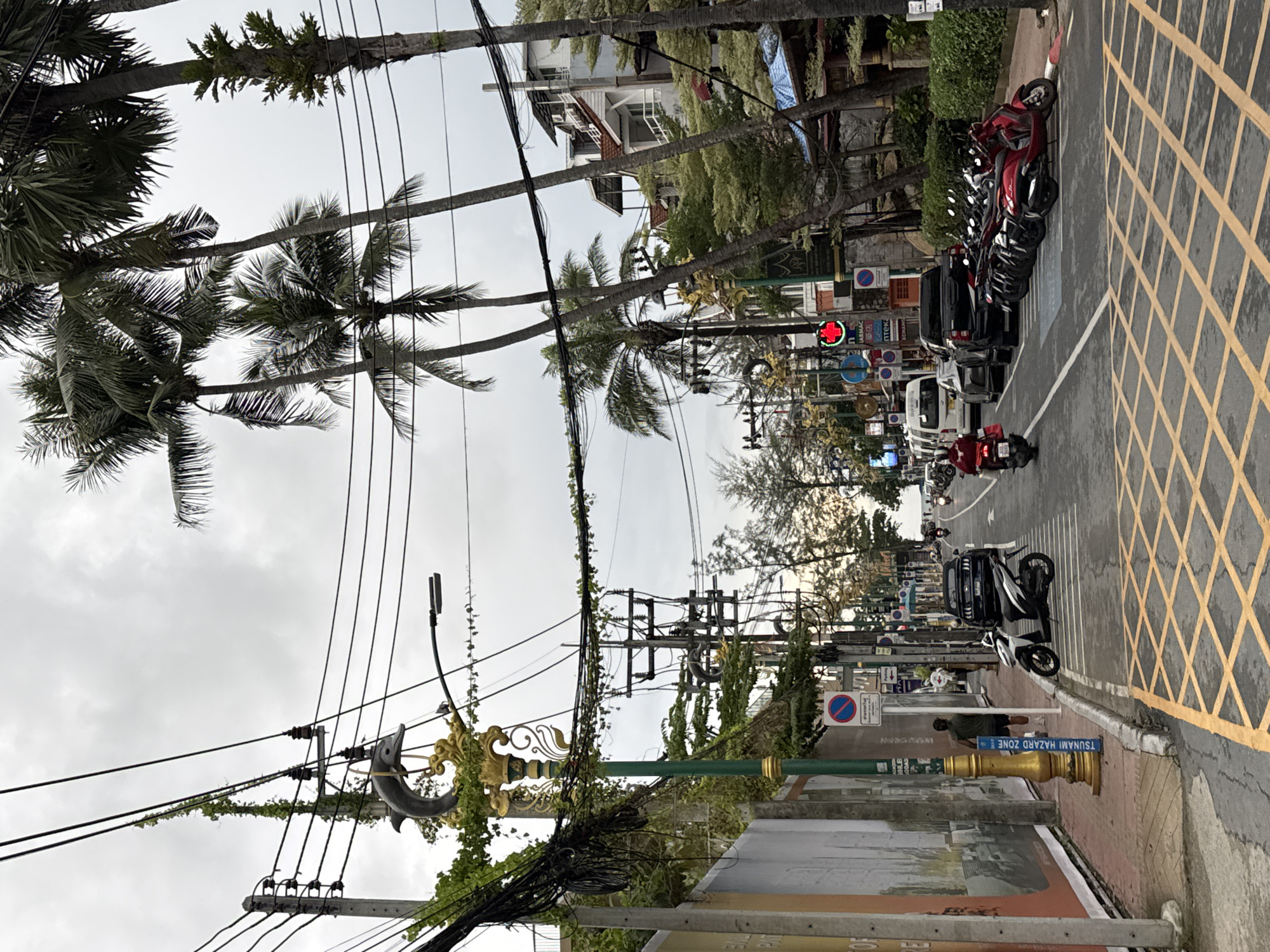 The image depicts a street scene in Patong, Thailand, with a cloudy sky overhead. On the left, a sidewalk runs alongside a building adorned with a large billboard and a decorative lamppost featuring a dolphin sculpture. The street itself is paved and marked with yellow diamond-shaped crosswalks and parallel white lines indicating parking spaces. A variety of vehicles are present, including several motorcycles and scooters parked along the right side of the road, a white van, a black pickup truck, and a grey car. Two individuals are visible: one person is walking on the sidewalk, and another is riding a red scooter. Palm trees and other vegetation line the street, and electrical wires are strung across poles. Buildings, some with multiple stories and balconies, are visible on the right side of the street. A traffic signal showing a red light is also present. Signs are posted along the sidewalk, including one with a no-parking symbol and another that reads "TSUNAMI HAZARD ZONE".