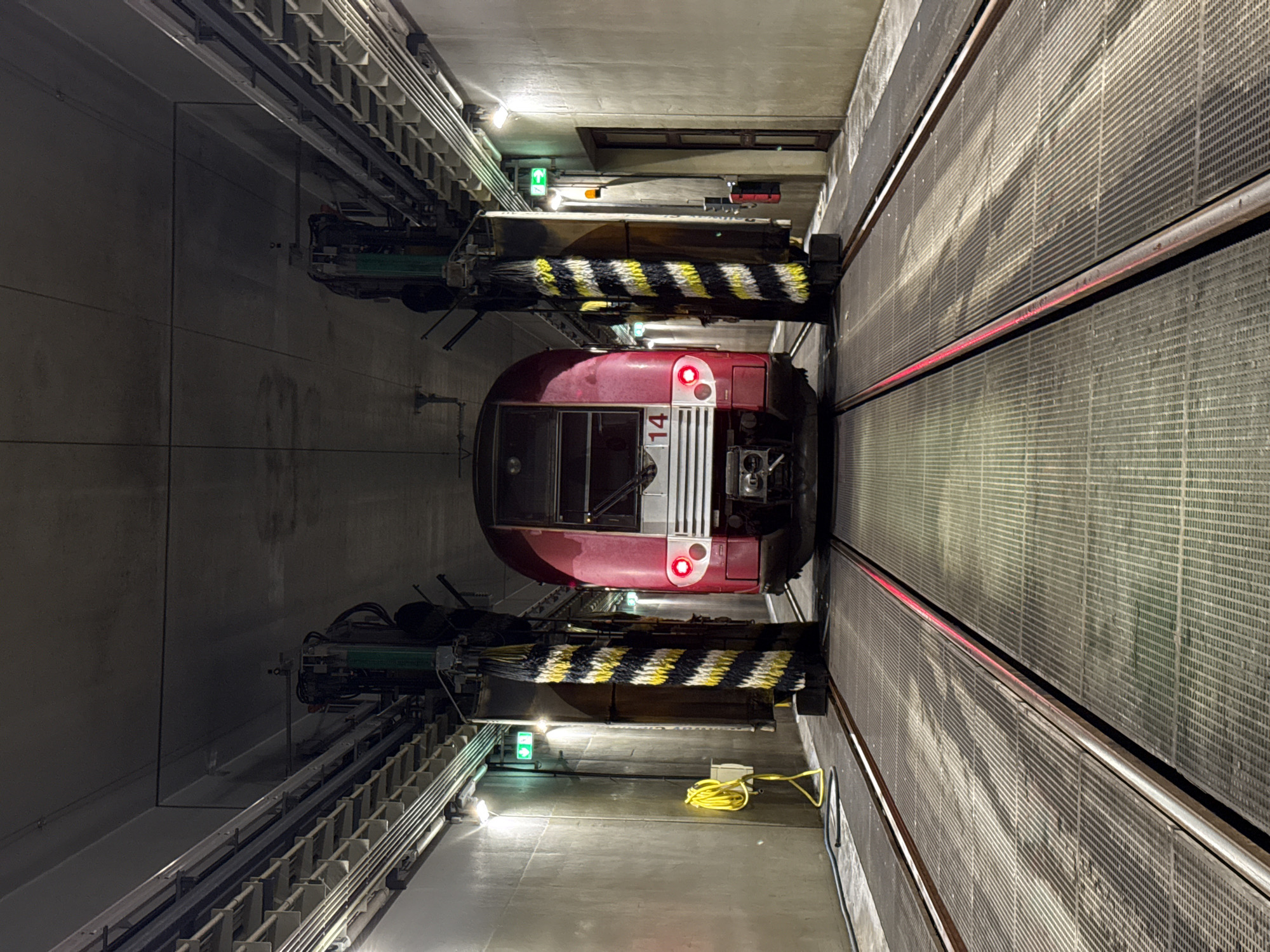 A red and white train car, number 14, sits centered in a narrow, illuminated tunnel. It's flanked by two train washing machines with yellow and black striped brushes. A coiled yellow cable rests on the tunnel floor. Green exit signs are visible on the ceiling above the washers. Gridded panels line the right wall, with a red light strip running horizontally. The tunnel is gray and appears to be concrete. The location is likely in Luxembourg, Luxembourg.