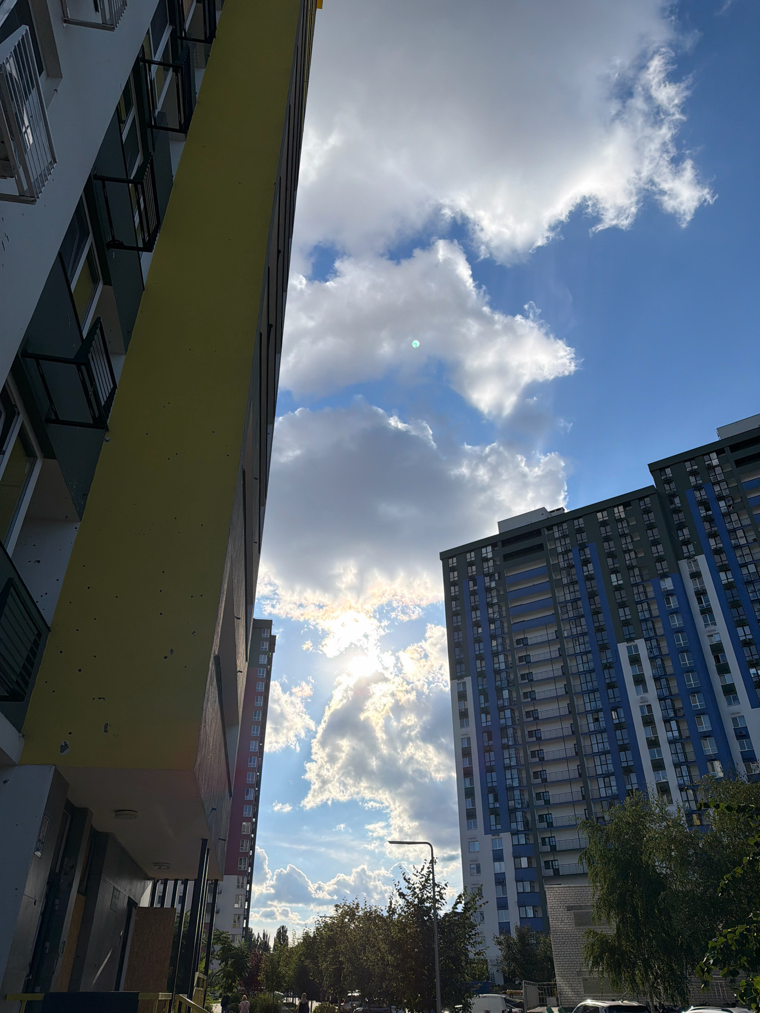 The image shows a street view in Kyiv, Ukraine, with two tall apartment buildings dominating the foreground. The building on the left is painted yellow and gray, with visible balconies and windows. The building on the right is taller and features a blue and gray facade with numerous windows and balconies. Sunlight streams through the clouds, creating a bright sky with some lens flare. Below the buildings, there are trees and a few individuals walking on the sidewalk. Cars are visible in the lower part of the frame. The scene suggests a residential area in the city.