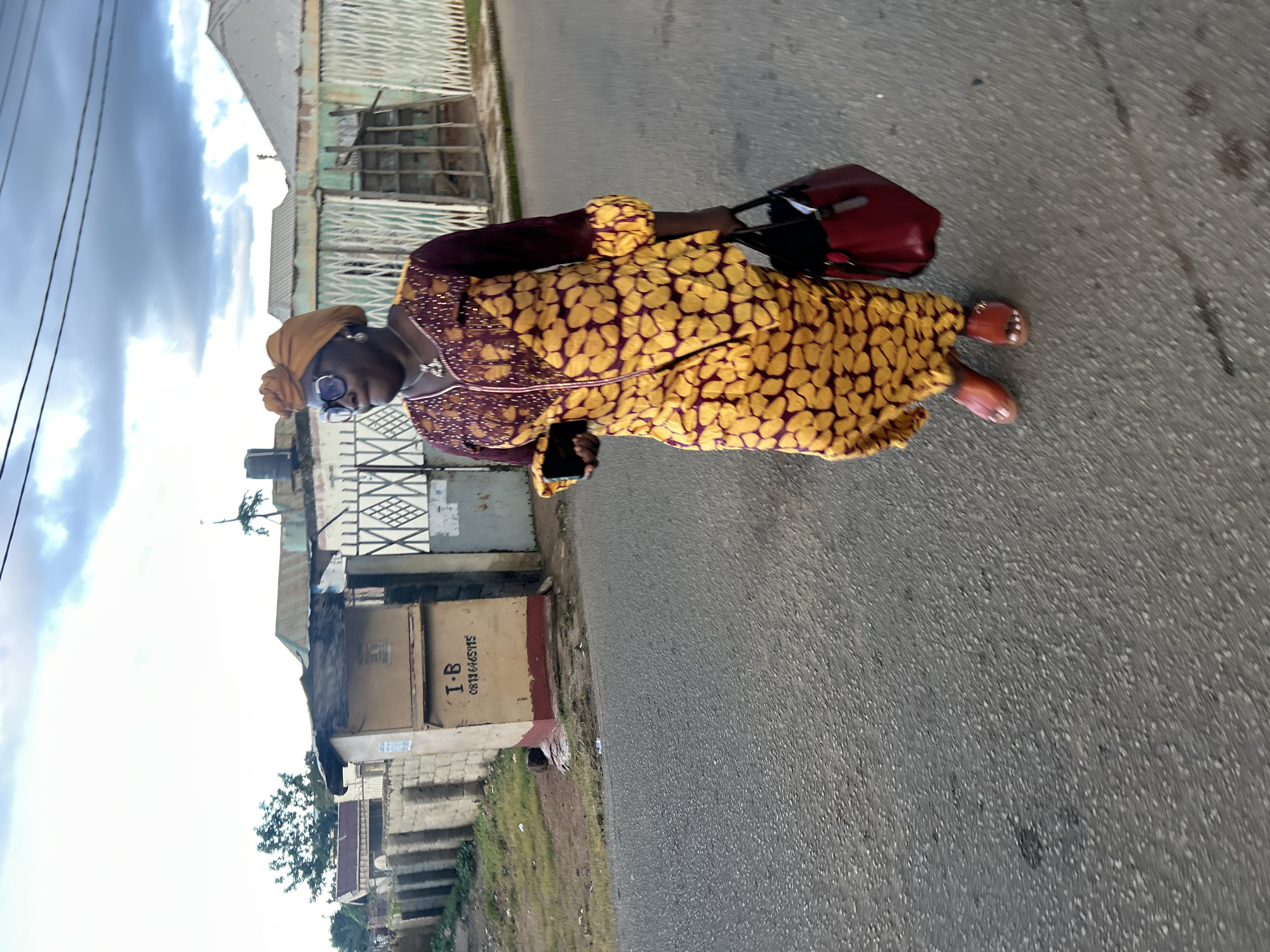 A woman is walking on a paved street in Jos, Nigeria, in front of some buildings. She is wearing a yellow and maroon patterned dress with a matching headwrap, sunglasses, and sandals. She is carrying a maroon handbag and a smartphone. The buildings in the background appear to be simple, one-story structures with corrugated metal roofs. There is a stone wall and a metal gate visible. The sky is cloudy.